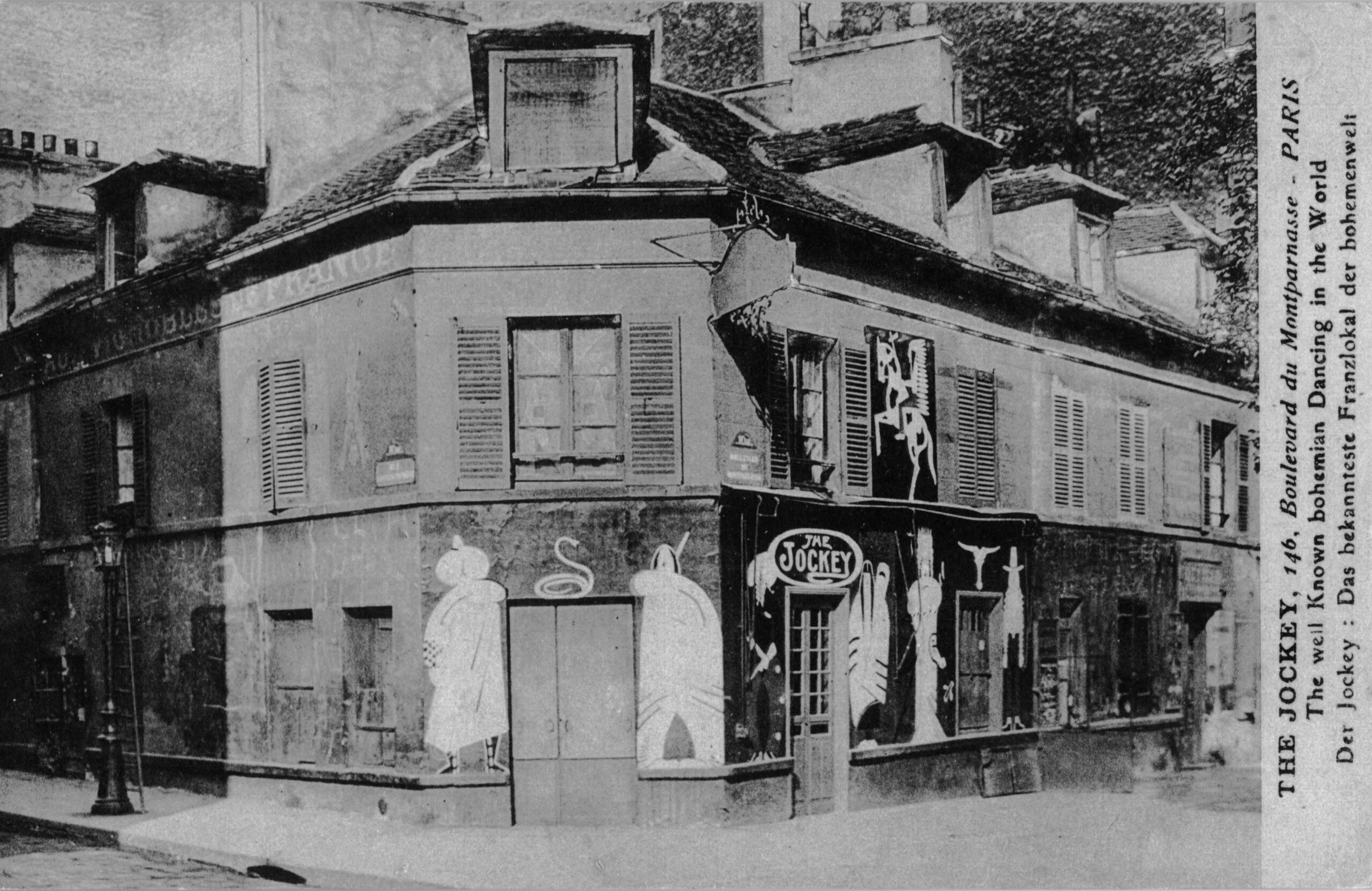 Black-and-white photograph of a corner building with murals painted on either side of a door way over which hangs a sign reading "The Jockey"