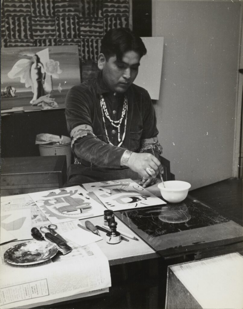 Black-and-white photo of a dark-haired man wearing beaded necklaces working on a painting