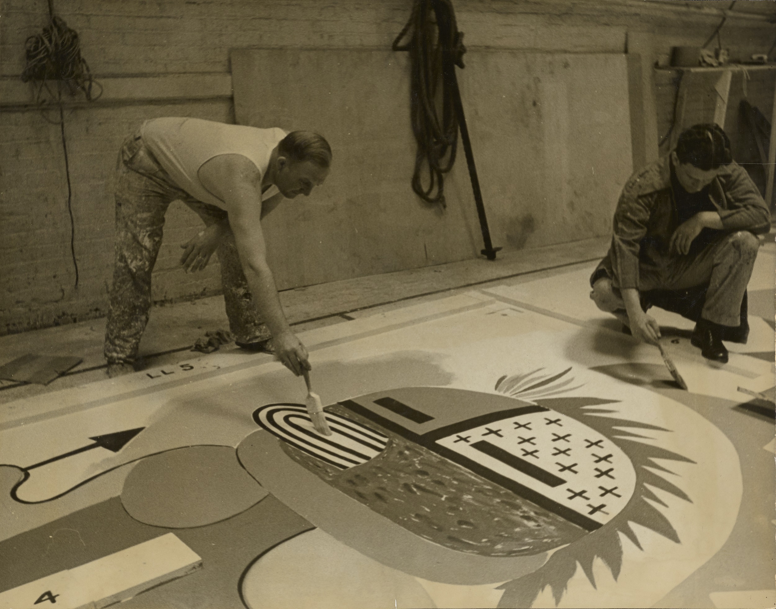 Black-and-white photograph of two men working on a painting, lying flat on the ground, in a Native American idiom