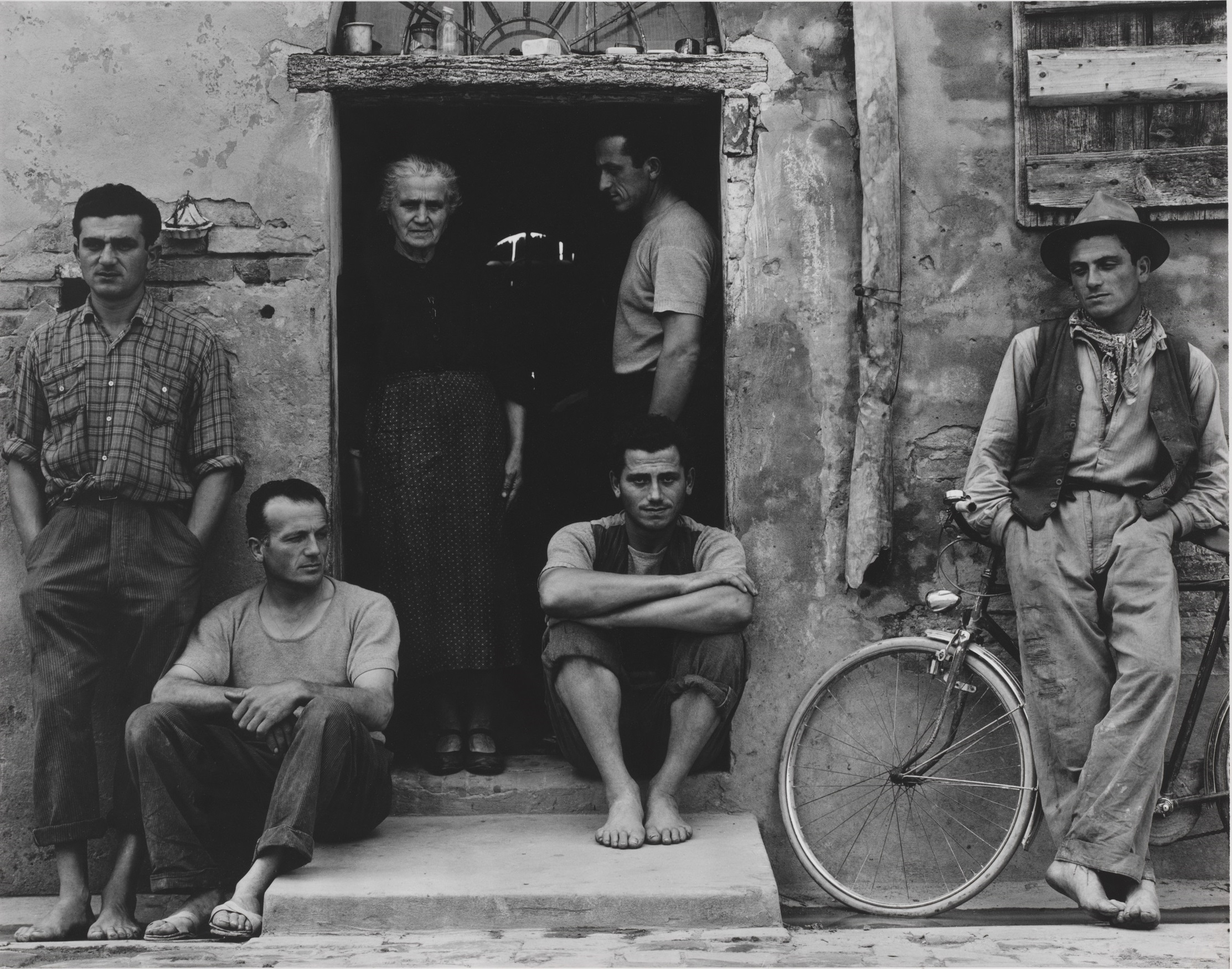 Black-and-white photograph of six Italian men seated outside the door way of a stone building. One of them men has a bicycle.
