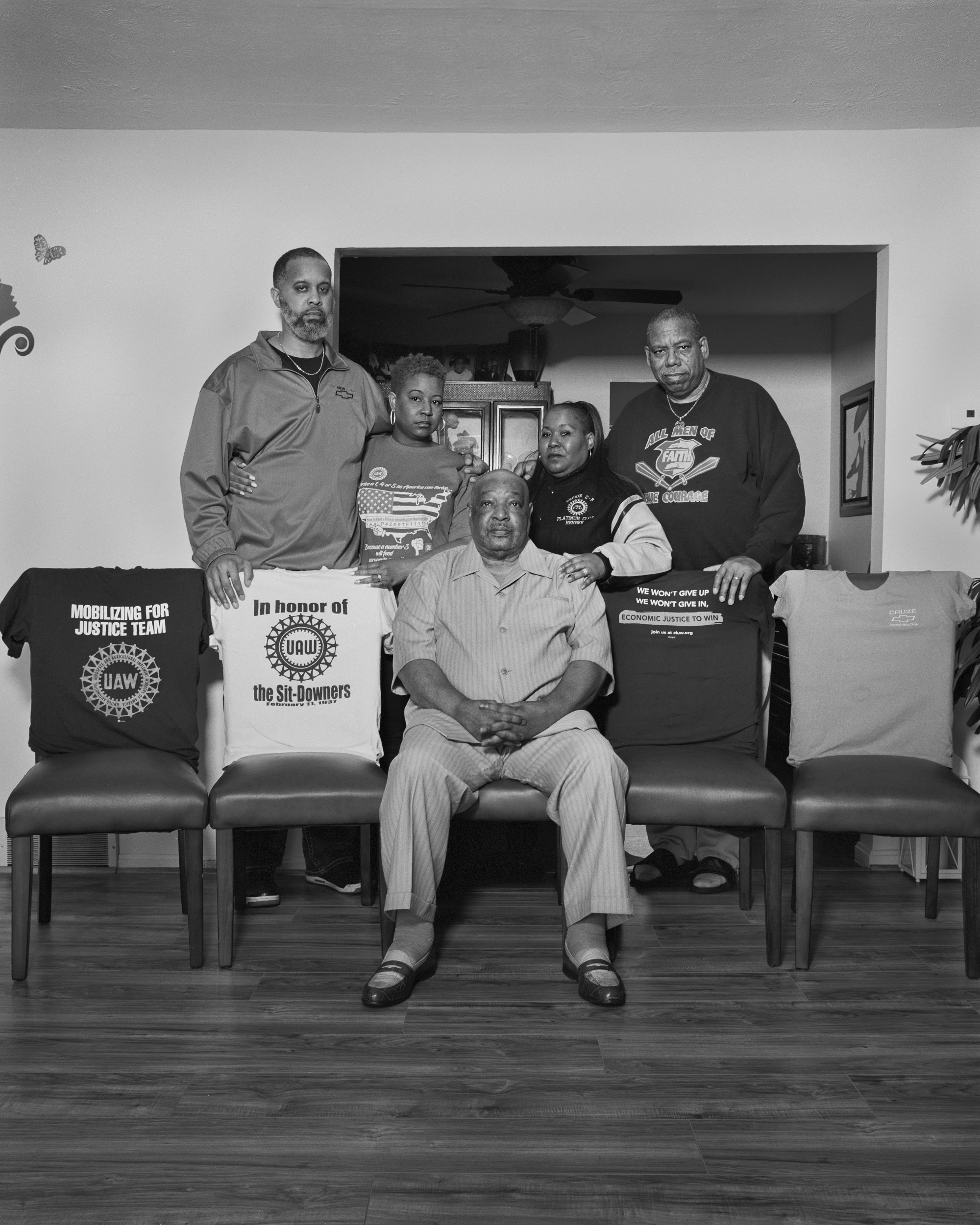 Black-and-white photograph of an African American family in a domestic interior. One man is seated in the center chair of a row of five chairs; the other chairs have t-shirts with pro-union slogans draped over the backs.