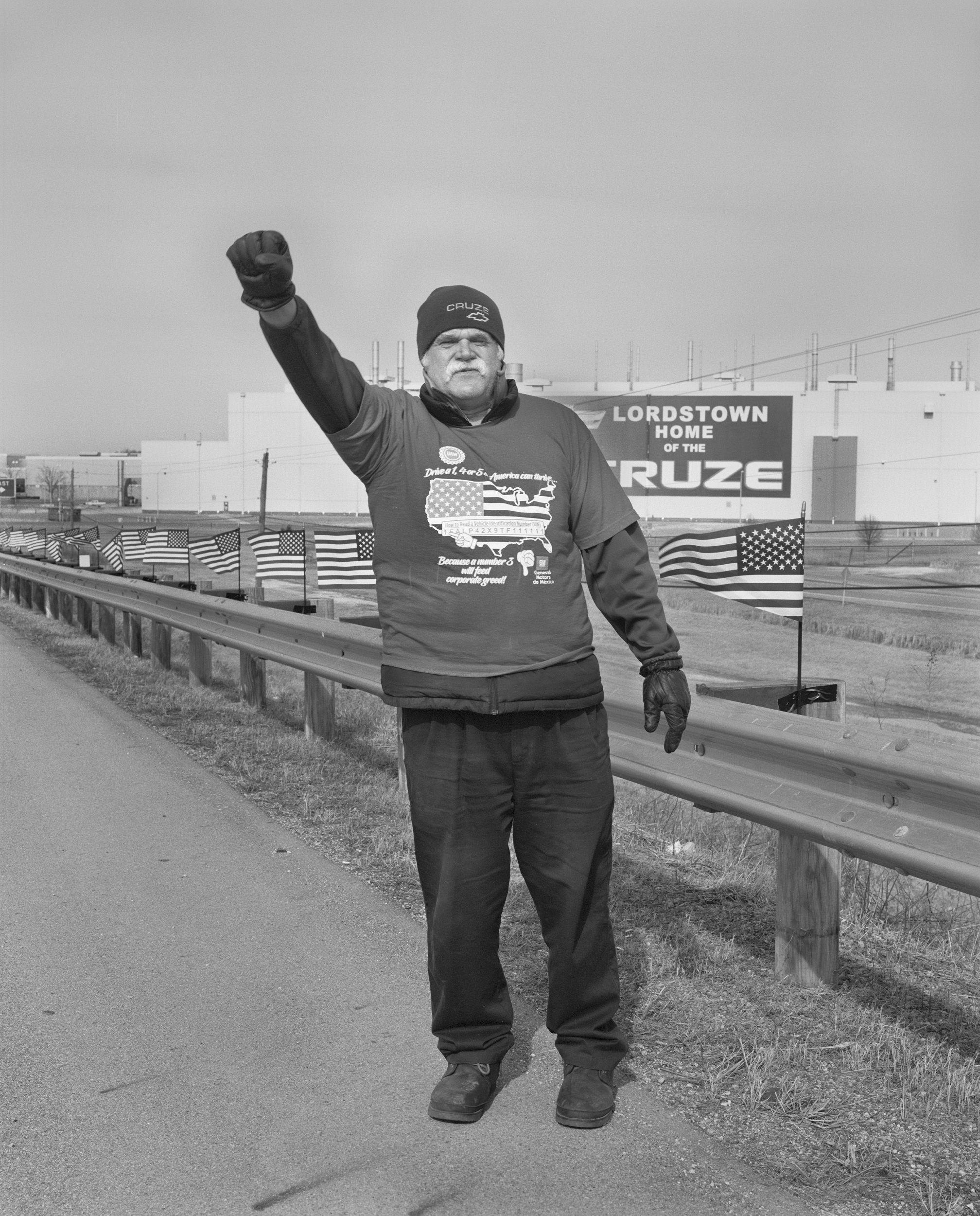 Black-and-white photograph of a middle-aged white man with a white beard standing in front of a sign reading "Lordstown Home of the Cruze" and raising his right fist to the sky.
