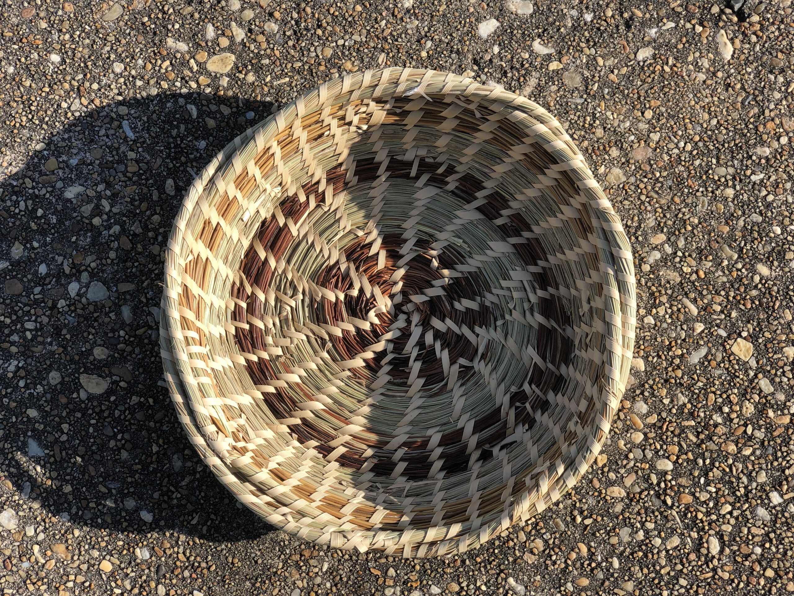 Coiled sweetgrass basket in different tones of buff, green, and brown, seen from above against an asphalt surface