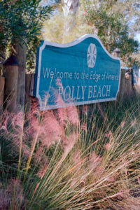 Feathery pink plantings in front of a blue-and white sign that reads "Welcome to the Edge of America FOLLY BEACH"