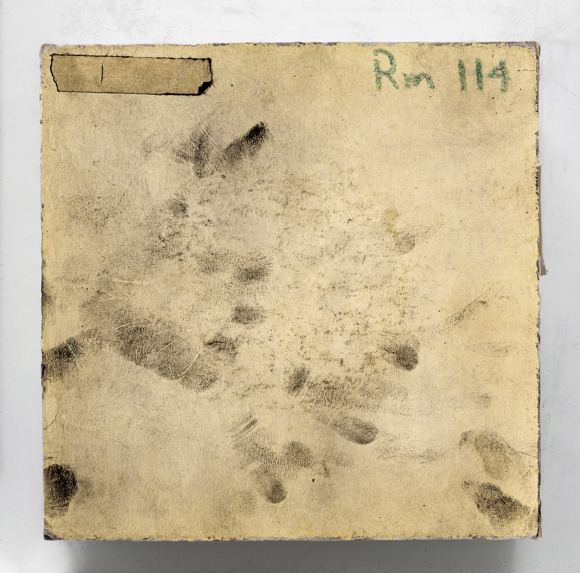 Buff-colored piece of paper covered with smudged black fingerprints. In the upper left corner is a piece of adhesive tape; in the upper rights, in green crayon, are the words "Rm 114" 