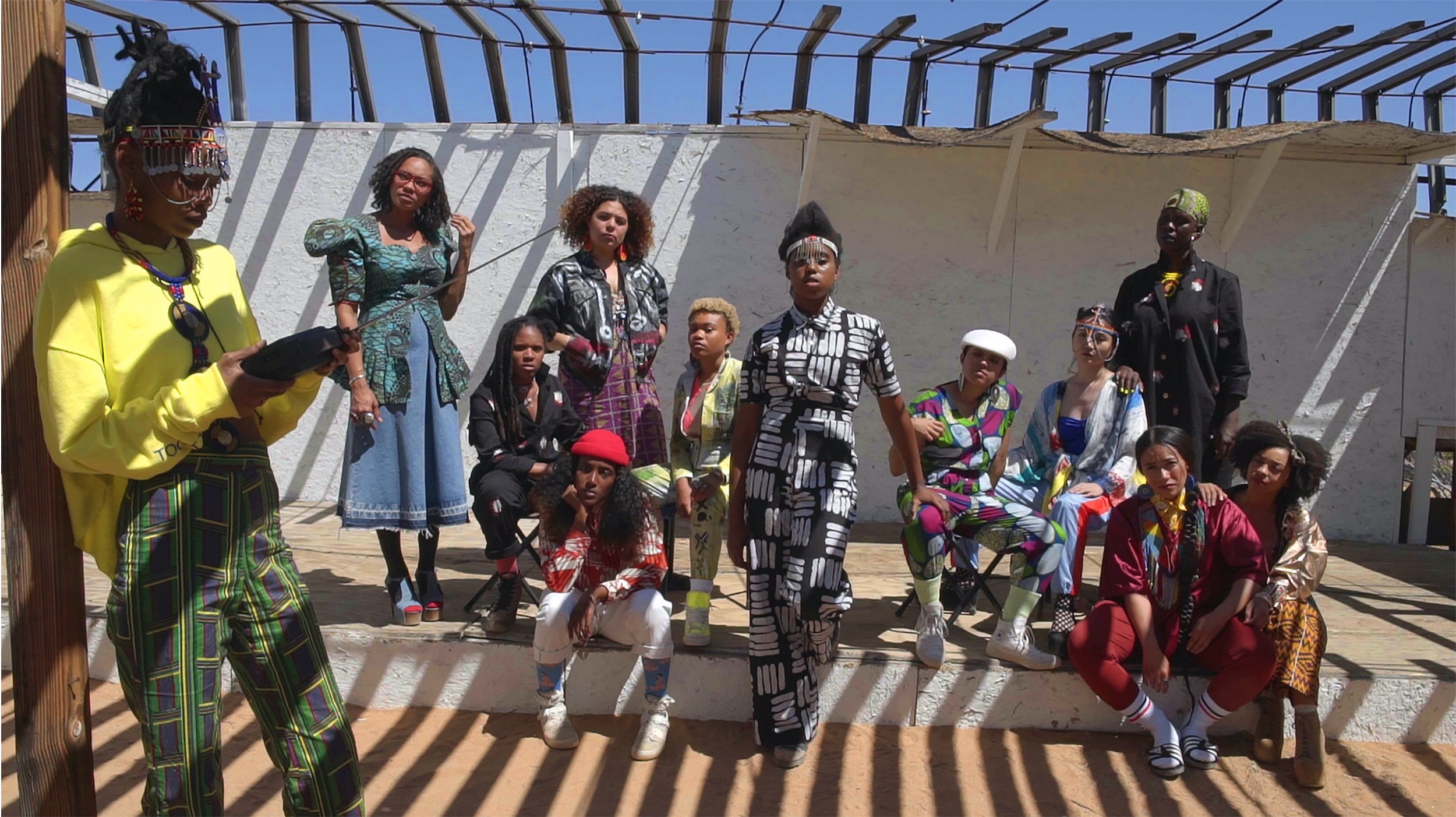 Color photograph of twelve young Black people in colorful clothing gathered outdoors under a pergola