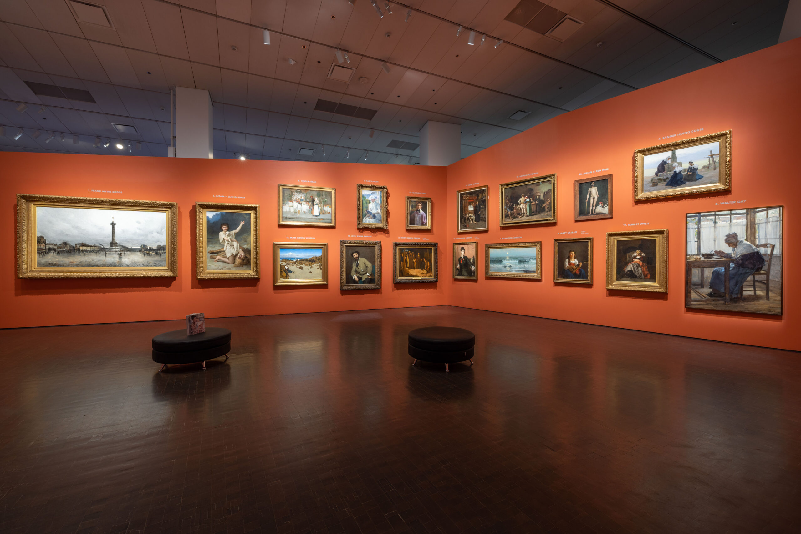 View of the interior of a museum gallery, with two temporary walls, painted orange, joined in a corner. Seventeen framed paintings are hung densely on the walls.