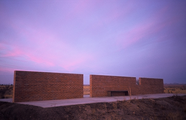 Sunset view of an outdoor sculpture consisting of a segmented brick wall on a concrete plinth in a desert landscape