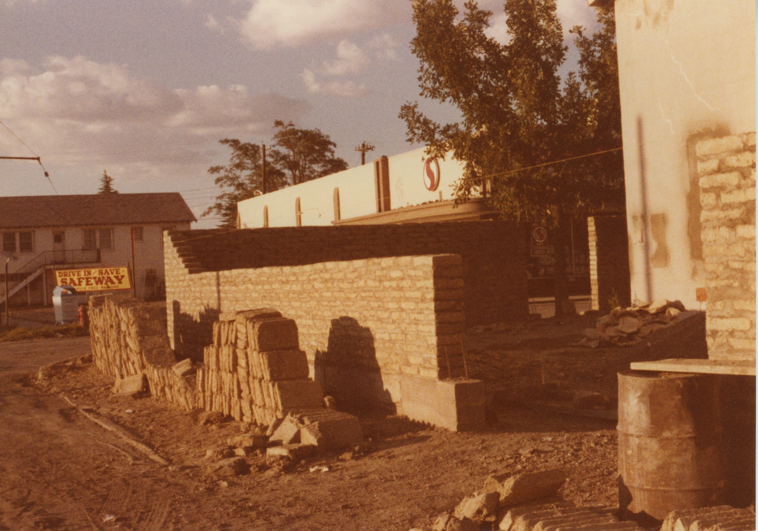 Historical color photograph of a street corner with a pile of adobe bricks. Just beyond a sign reads "Drive In Save / Safeway"