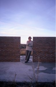 Color photograph of a person standing within an opening between two brick walls, leaning to their left