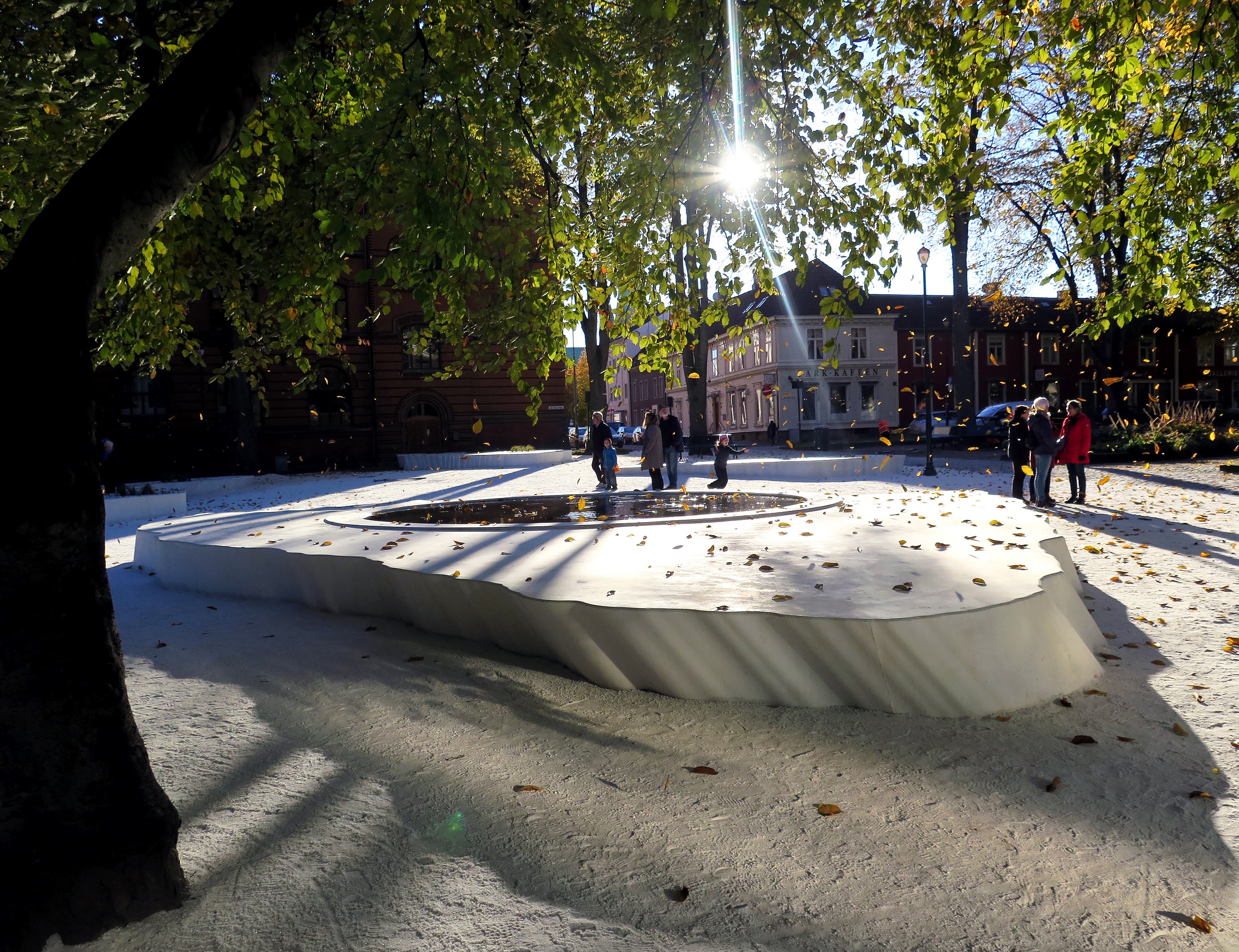 Sunlit view of a park with an outdoor sculpture consisting of an irregular flat form about a half-meter high, with an aperture in the middle