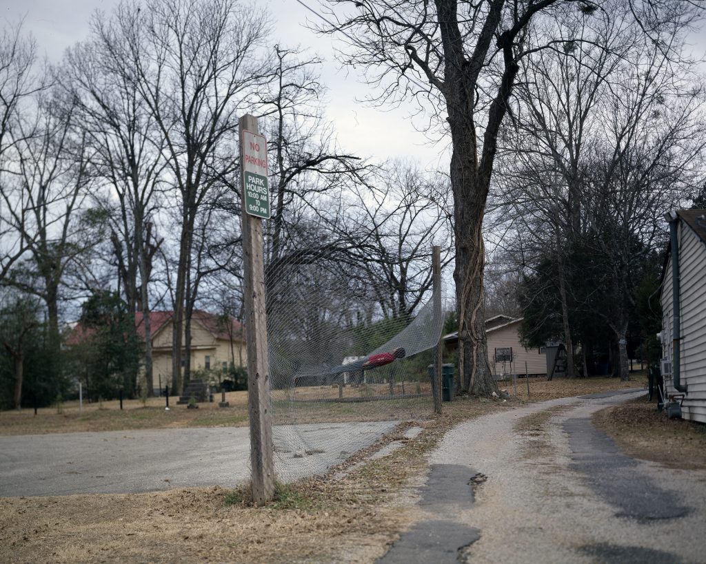 Color photograph of a working-class street corner, with a boy in a red shirt resting in a net between two poles. Signs on the nearest pole read "NO PARKING" and "PARK HOURS 10:00 AM TO 8:00 PM"