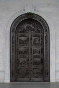 Set of large bronze doors with carved reliefs and a curved top set into a marble wall.