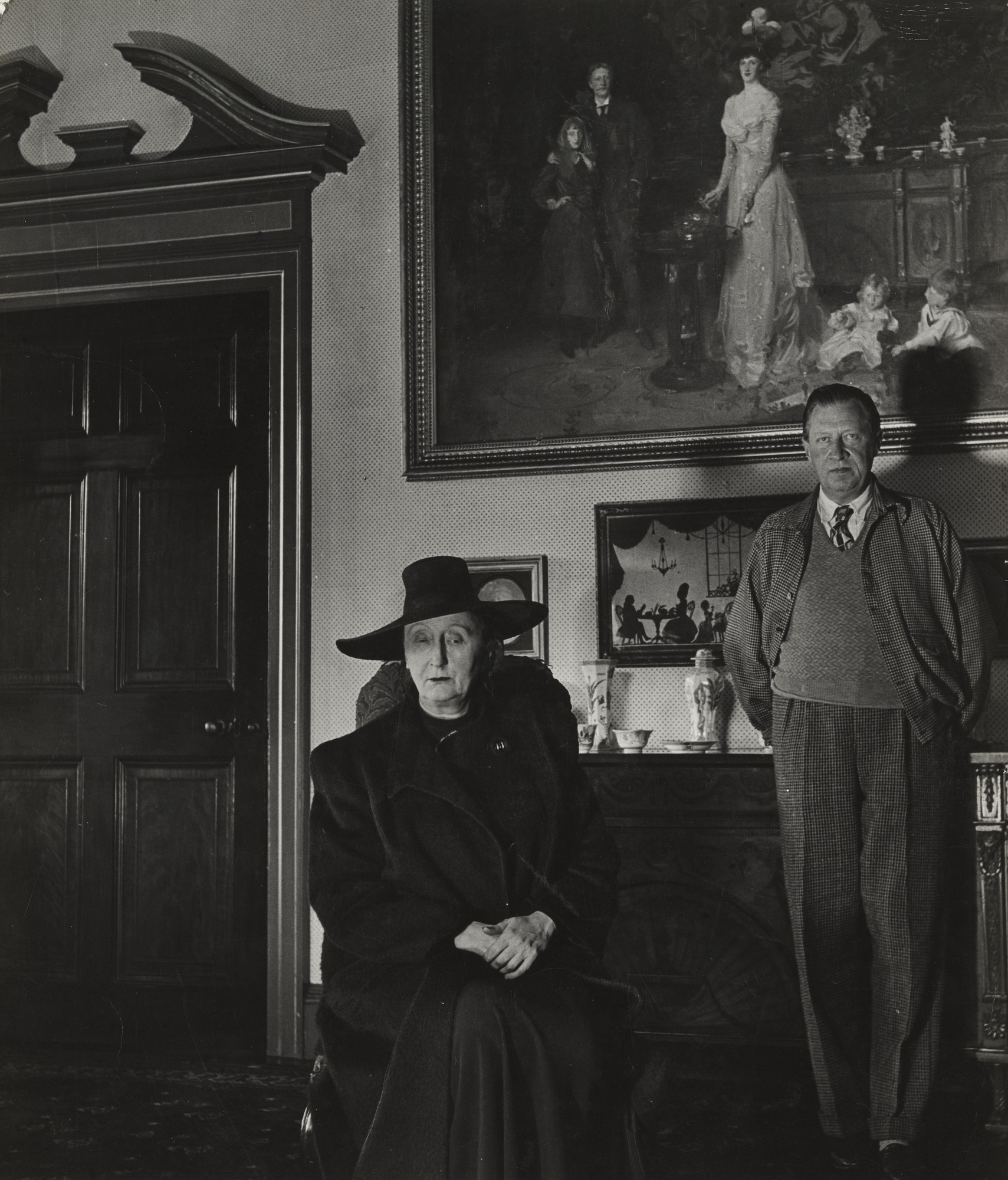 Black and white photograph of a seated woman in a black hat and a standing man in a sweater and tie in front of the painting in fig. 2.