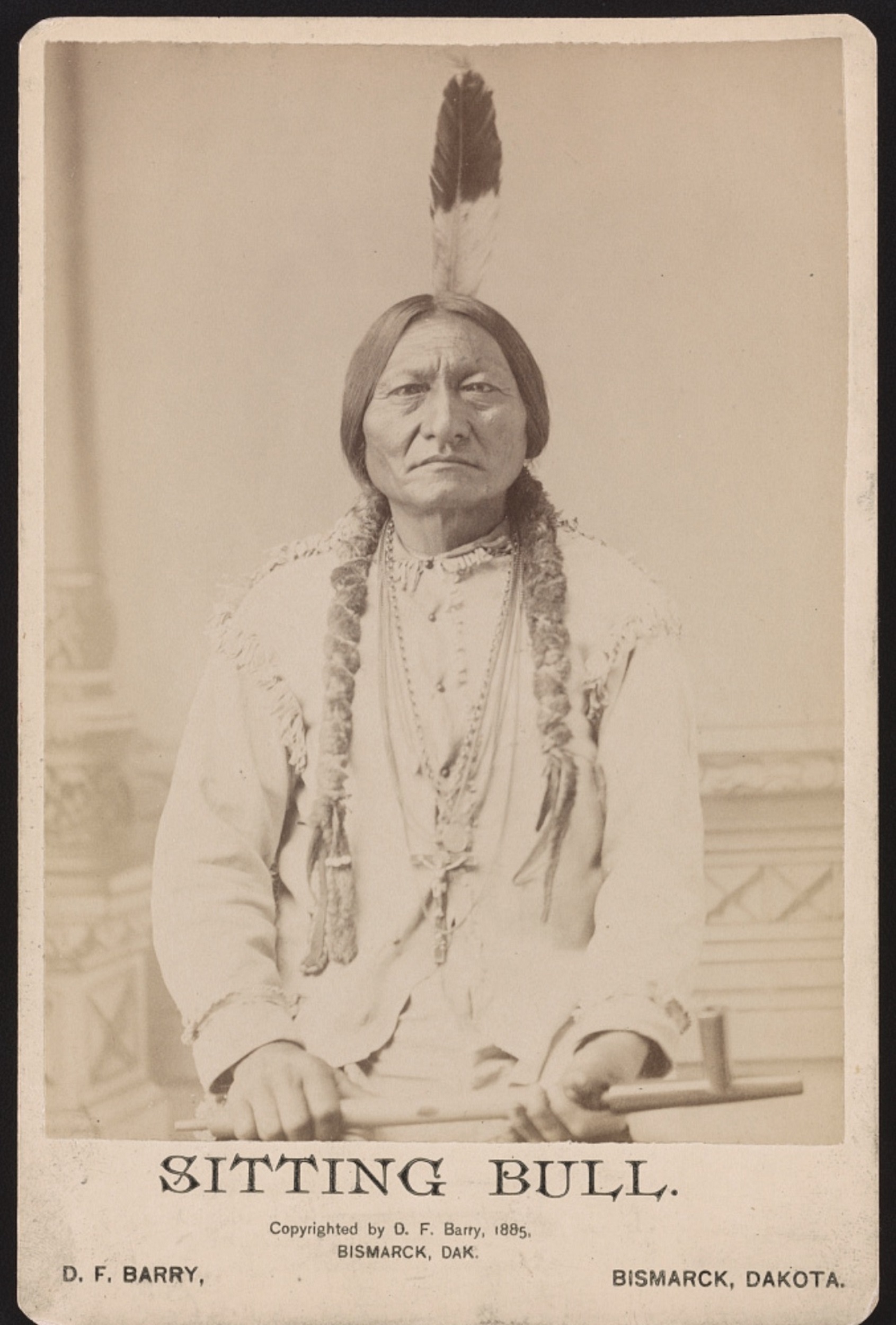 Sepia-tone carte de visite photograph of a Native American man with a tall father on his head. The words below read "Sitting Bull / Copyrighted by D. F. Barry, 1885 / Bismarck, DAK. / D. F. Barry / Bismarck, Dakota