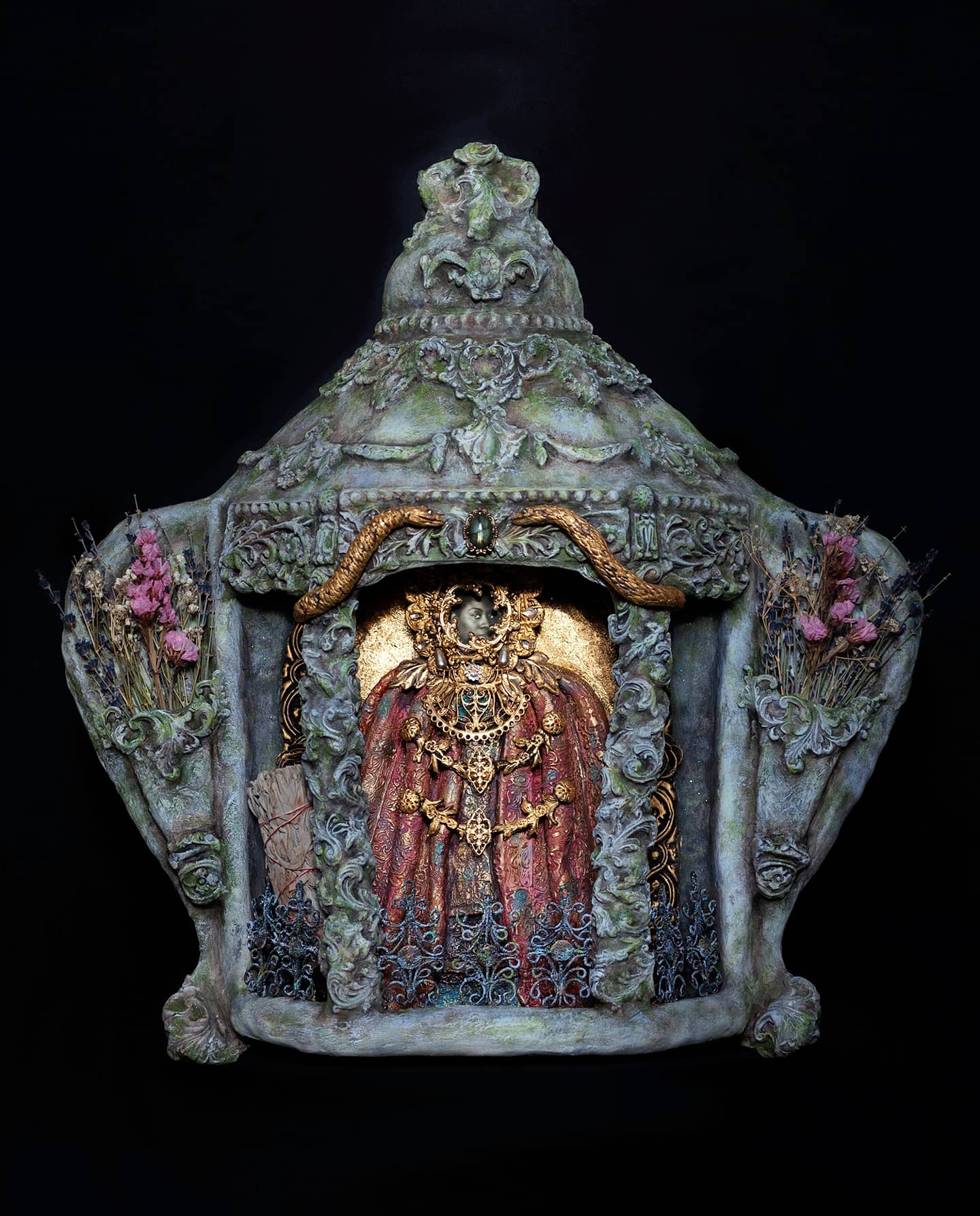 An ornate greenish-gray object with a pointed top set against a black background. The front of the object has a figure rendered in gilt, wearing a red cloak.