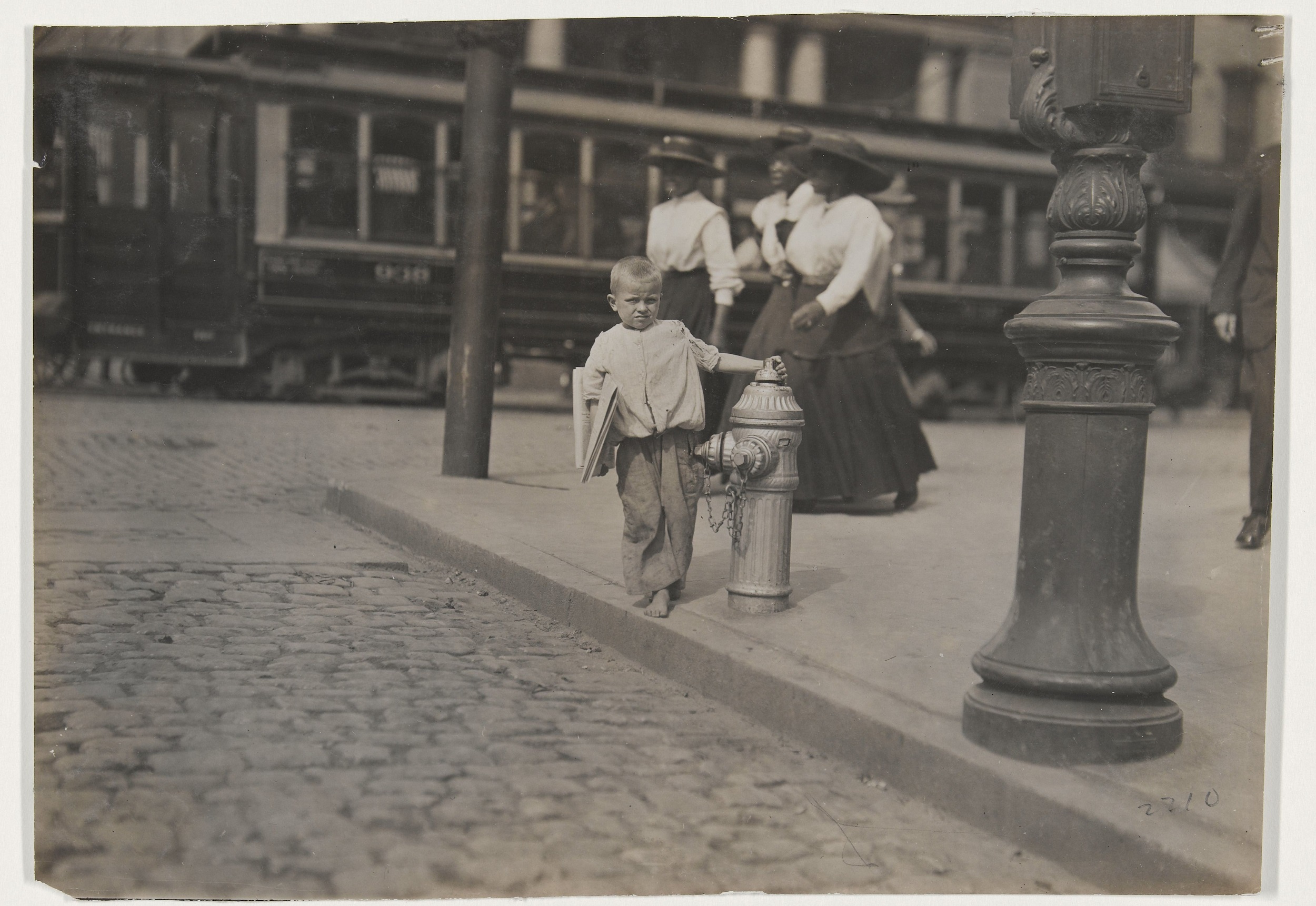 Black-and-white photograph of a young blond boy with newspapers under his arm, standing on a street corner. In the background, three Black women in hats, white shirts, and dark skirts prepare to cross the street.