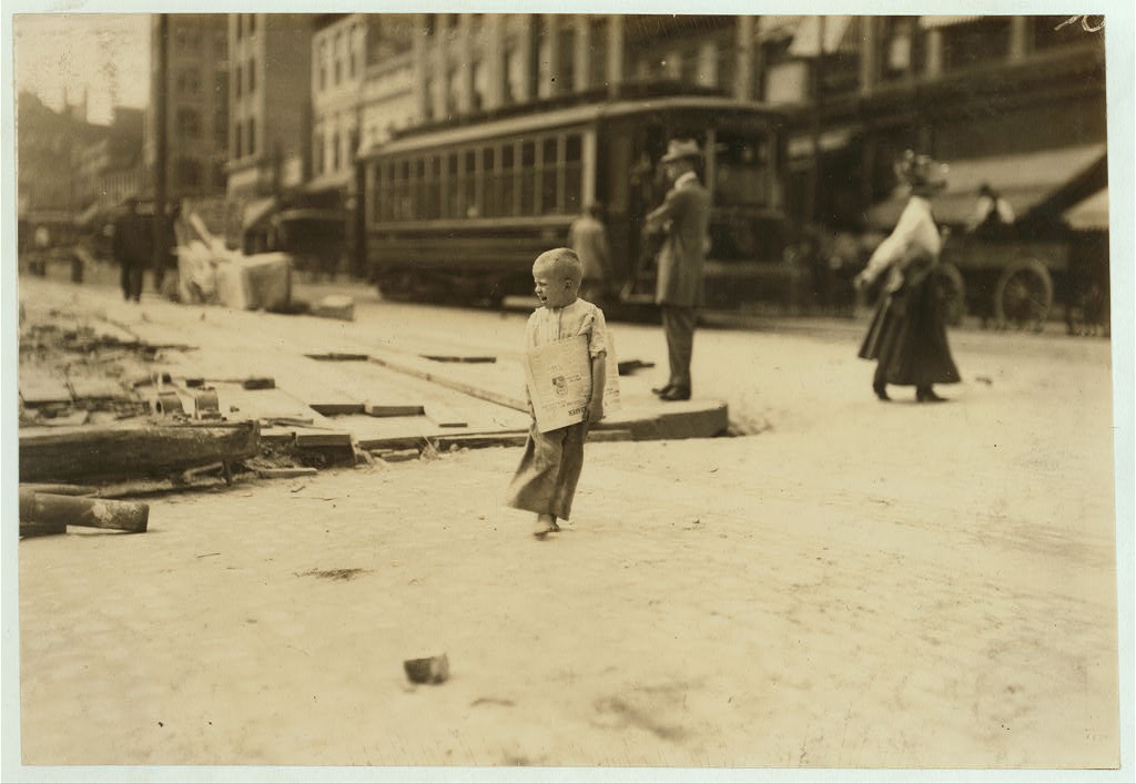 Sepia-toned black-and-white photograph of a barefoot young blond boy with a newspaper under his arm crossing the street. behind him are a well-dressed man and woman and a trolley car.
