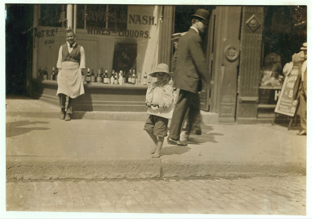 Sepia-toned black-and-white photograph of a barefoot young white boy in a hat, holding a newspaper and stepping down from a curb. Behind him is a dark-skinned man in a white apron standing in front of a storefront, and another man in a dark suit and bowler hat.