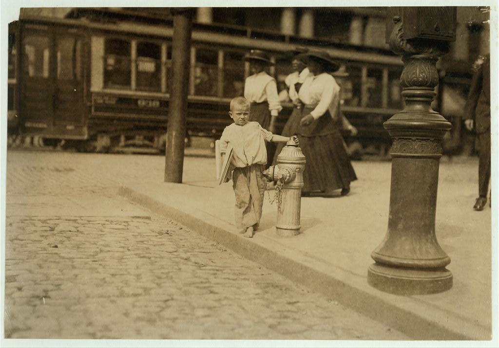 Sepia-toned black-and-white photograph of a young blond boy with newspapers under his arm, standing on a street corner (same composition as fig. 2). In the background, three Black women in hats, white shirts, and dark skirts prepare to cross the street.
