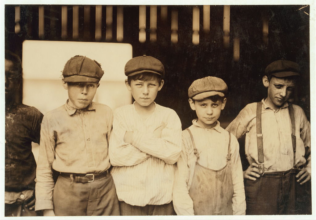 Sepia-toned photograph of five young adolescent boys wearing newsboy caps. The dark-skinned boy on the left is partially cut out of the frame.