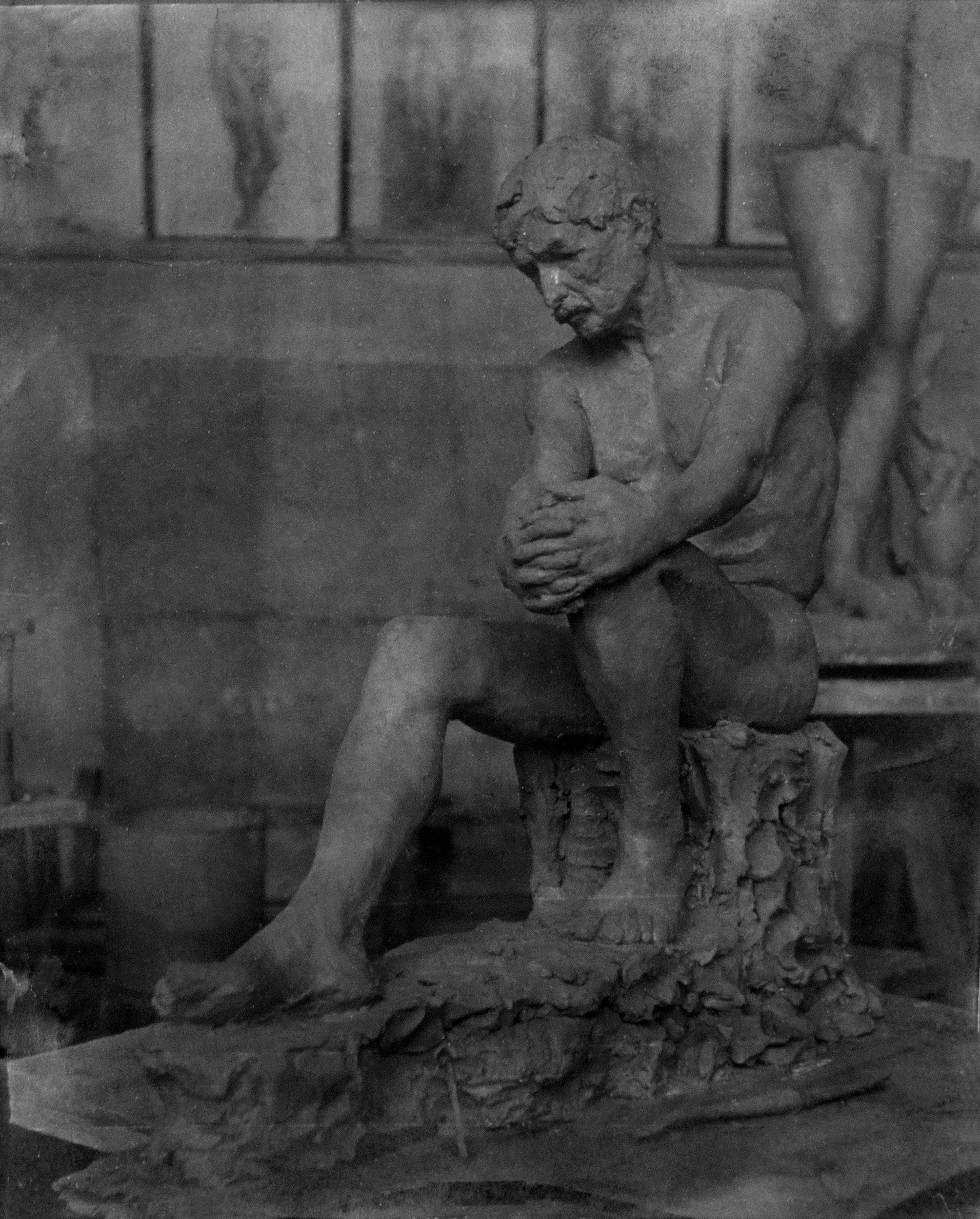 Historical black-and-white photograph of a sculpture in the form of a seated man, hands clasped over one knee