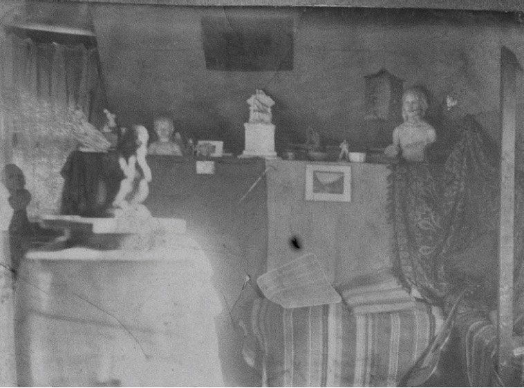 Historical black-and-white photograph of the interior of a sculptor's studio, with a striped couch and cushions in the foreground and several bust-length sculptures behind it.