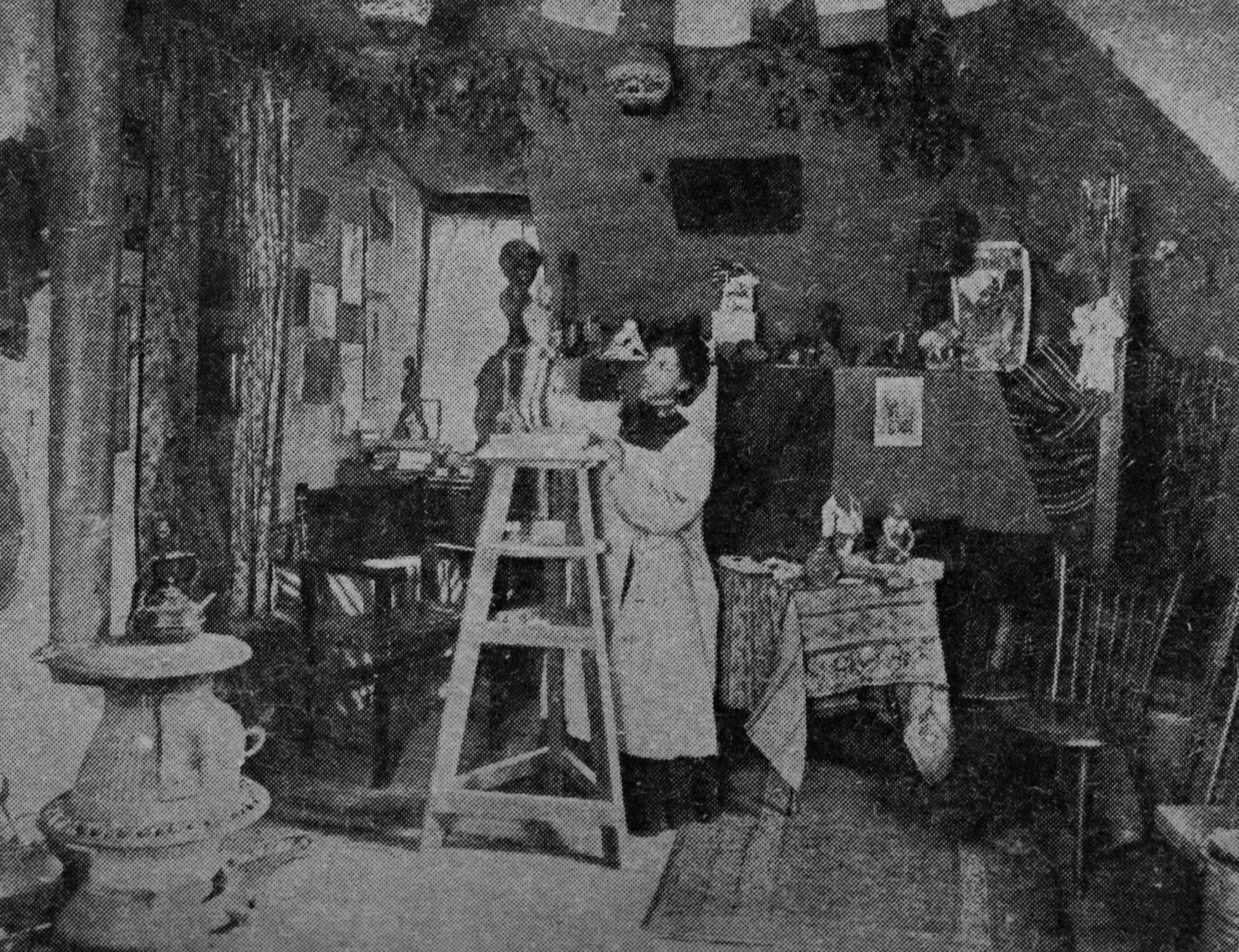 Black-and-white duotone photograph of a sculptor in her studio, working on a small sculpture on a wooden pedestal