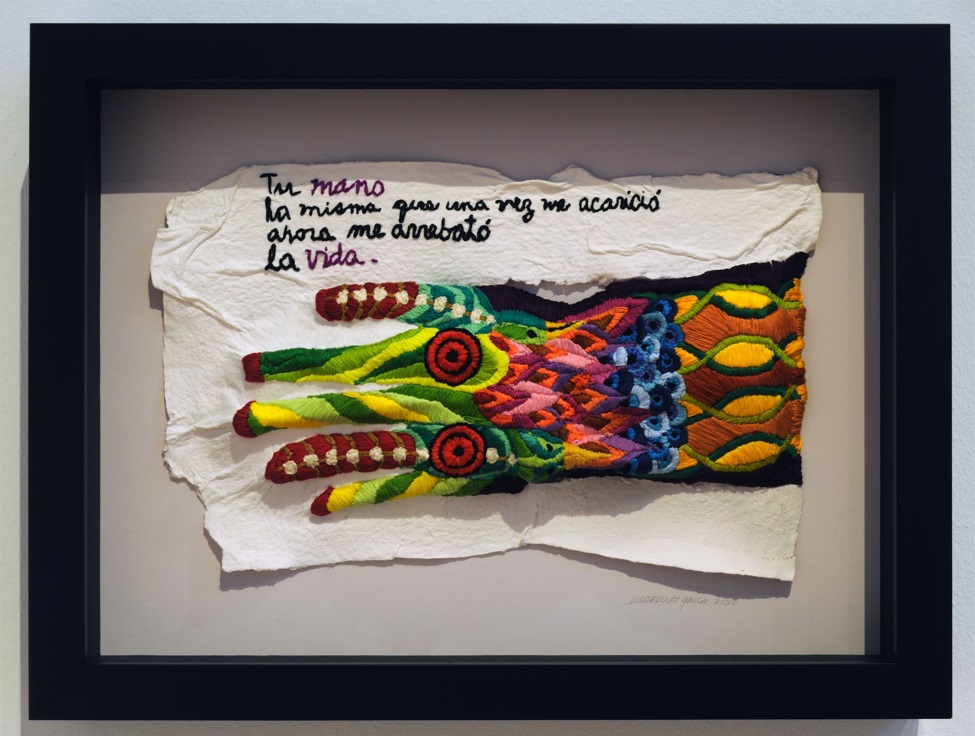 Framed work of textile art consisting of a colorful, embroidered hand or glove with these words above: "Tu mano / la misma que una vez me acarico / ahora me arrebato / la vida.'