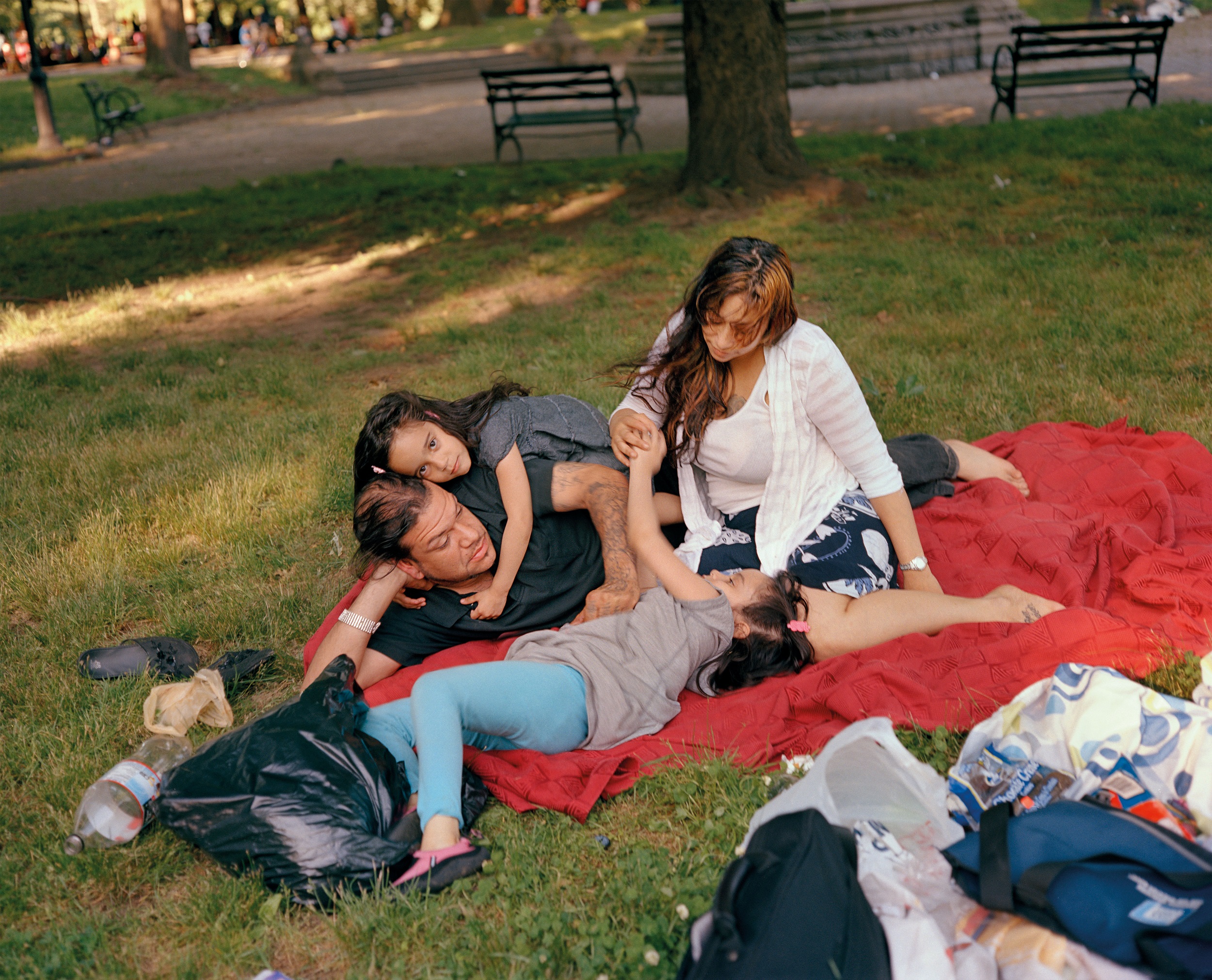 Color photograph of a man, woman, and two children picnicking on a red blanket in a park with grass, trees, and benches