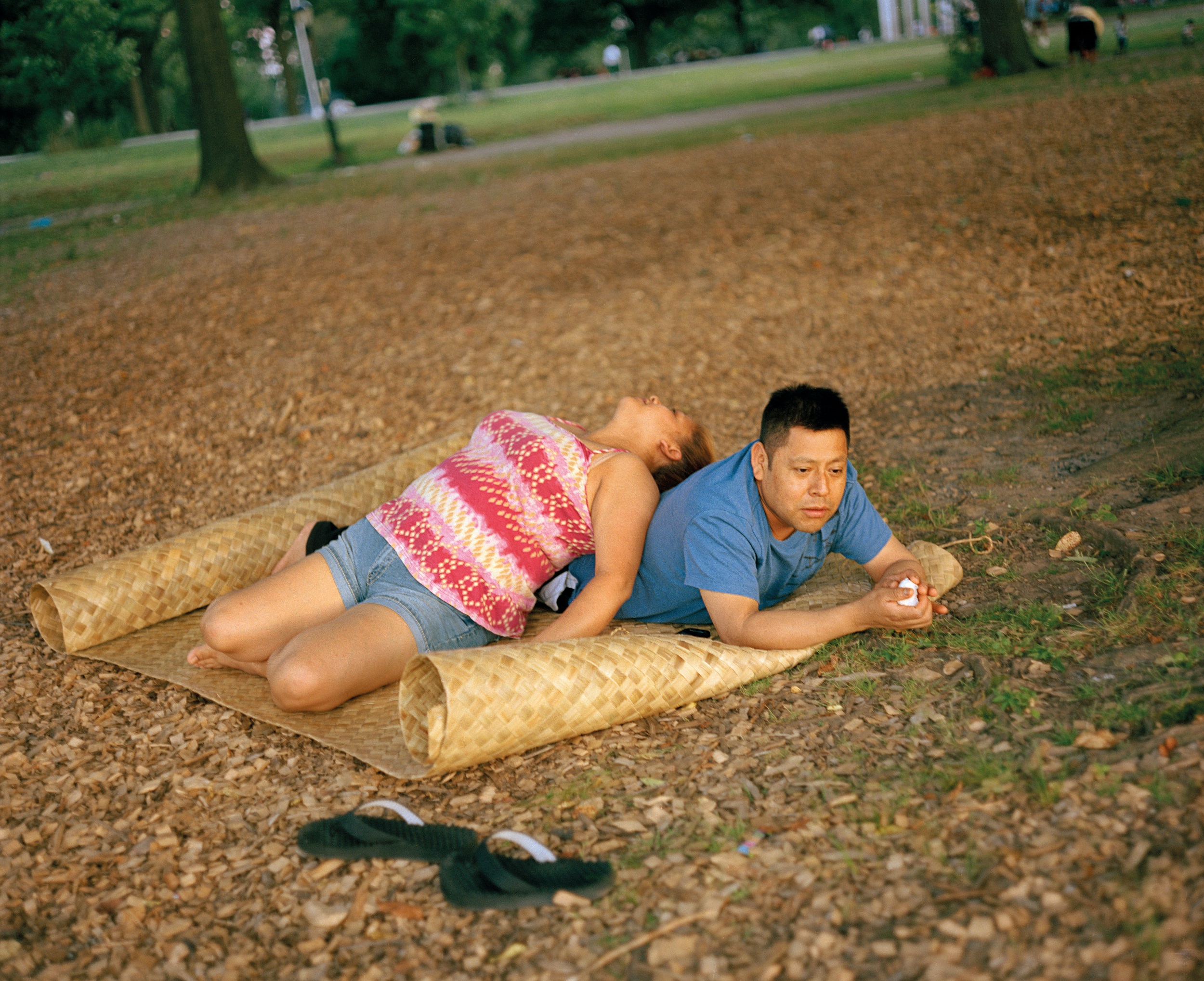 Color photograph of a man and a woman lyinig on a woven mat in a park. The woman is lying on top of the man and her legs are folded underneath her; her flip flops are in front.
