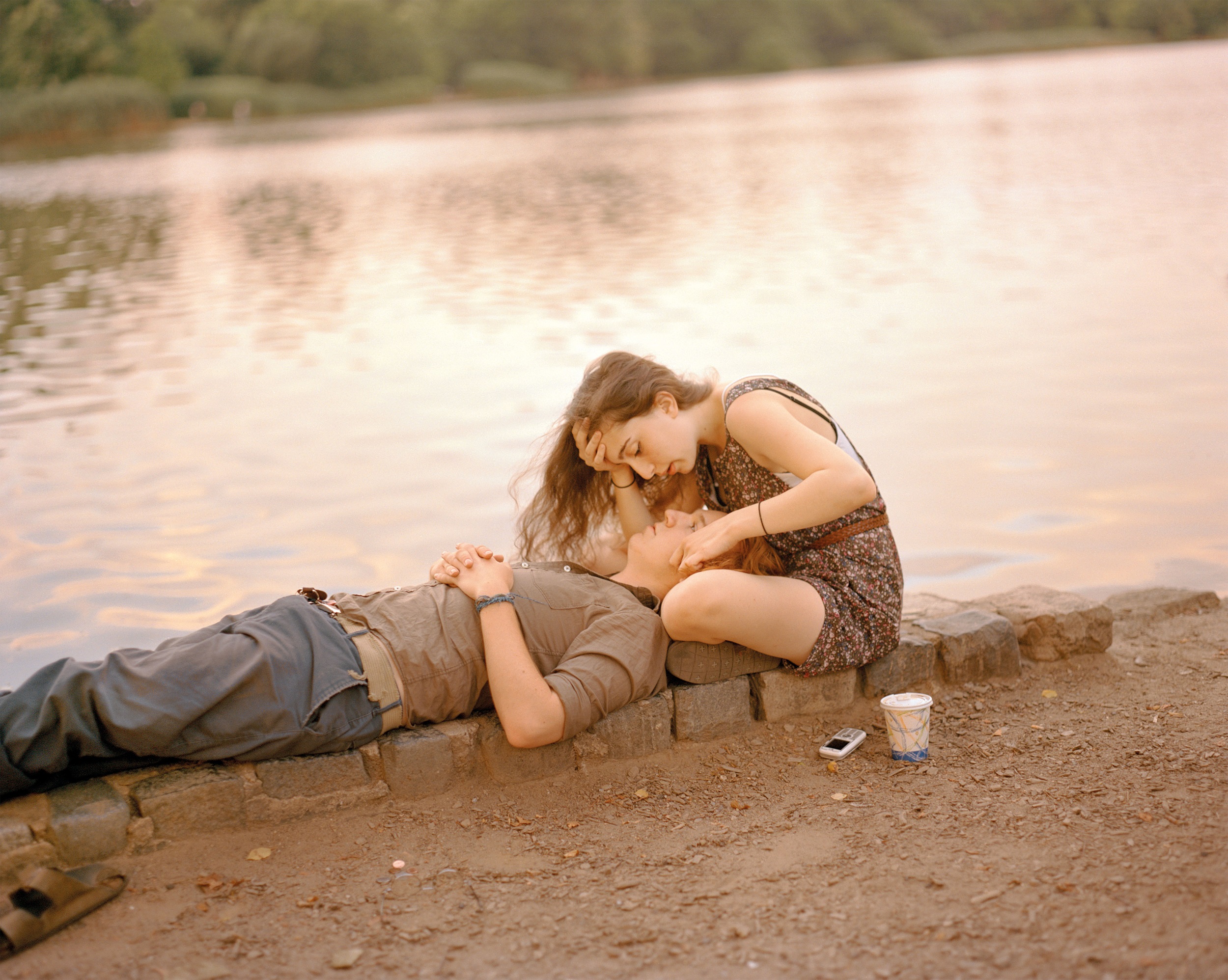Color photograph of a young couple by the edge of a body of water. The man is lying down with his head int he woman's lap; beside her is a disposable coffee cup and a cell phone.