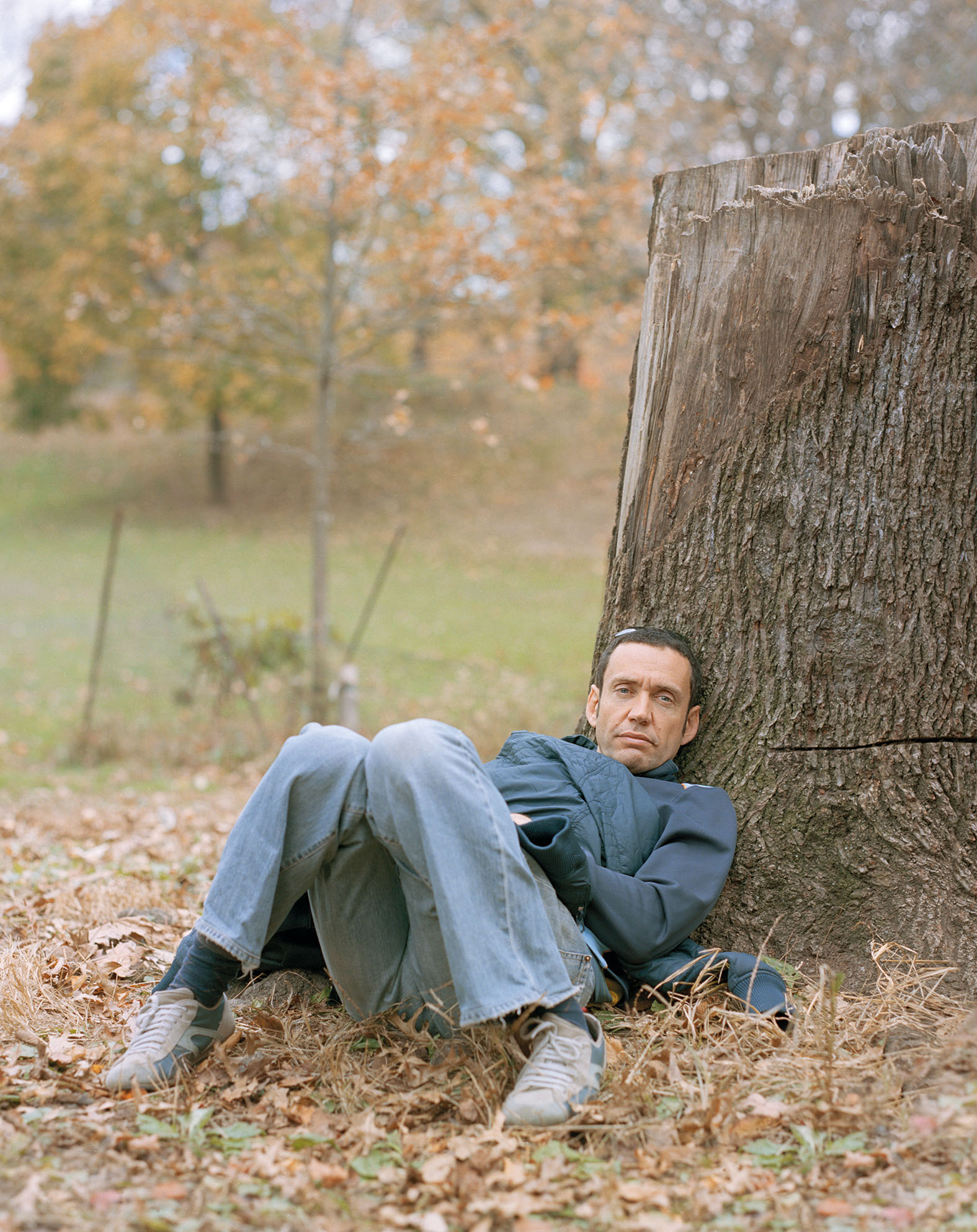 Color photograph of a man in blue jeans and a blue jacket slumped at the base of a tree stump in an autumnal landscape