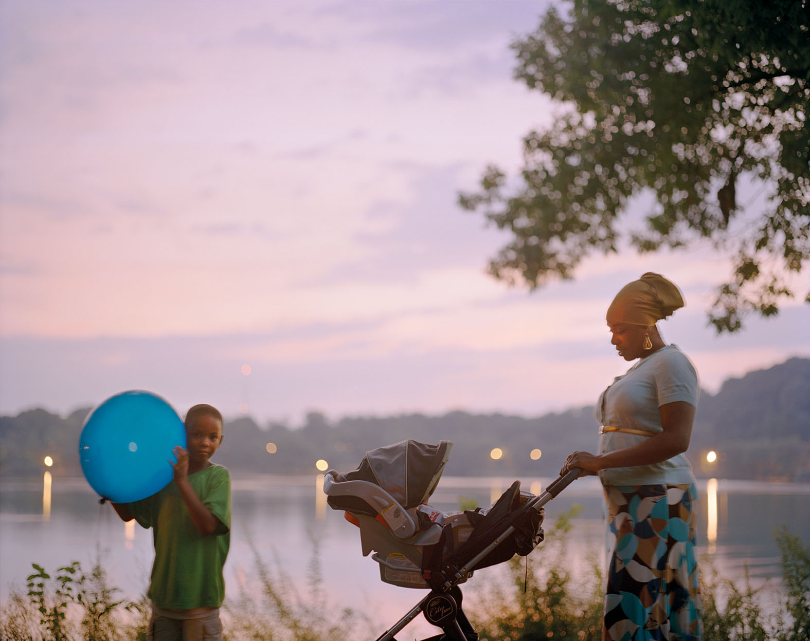 Color photograph of a woman pushing a stroller in front of a sunset sky over a body of water. To her right is a young boy holding blue ball; lights from the opposite bank are reflected in the water. 