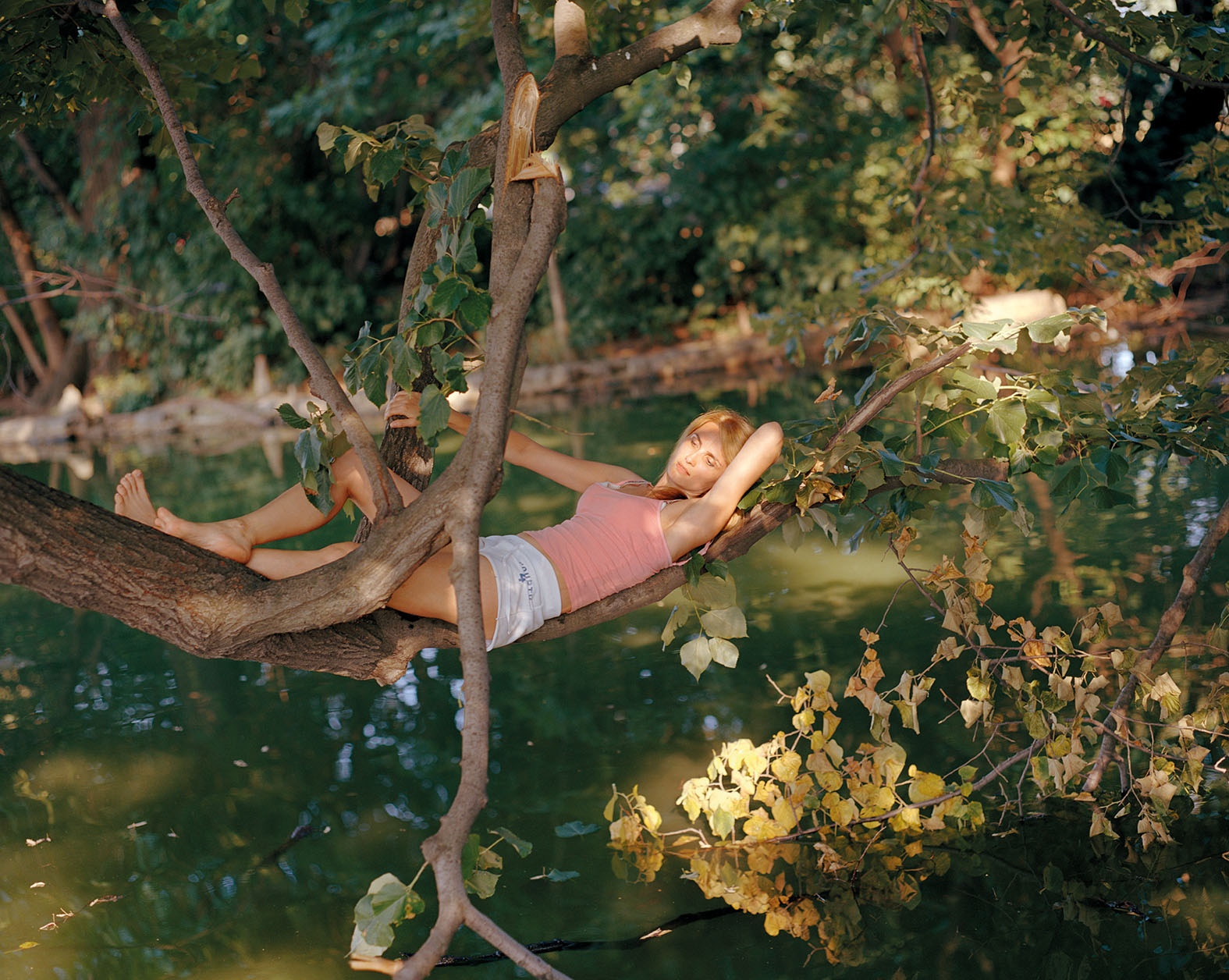 Color photograph of a blonde woman in white shorts and a coral-colored tank top resting in the branches of a tree.