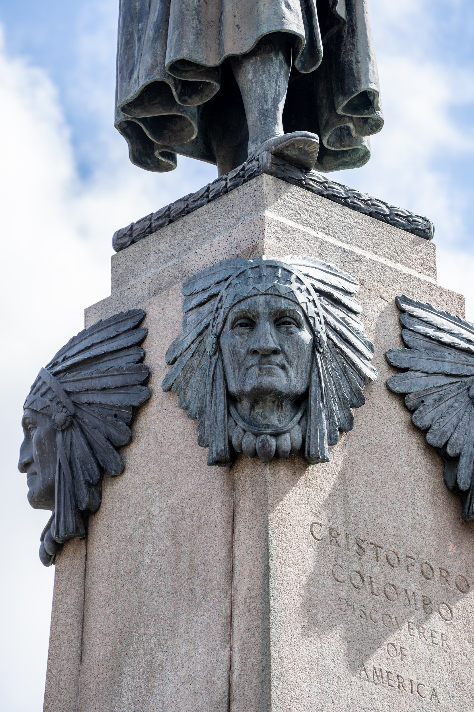 Detail of the base of the monument in fig. 3, showing bronze heads of Native Americans, wearing Plains war bonnets, attached to the corners of the pedestal, below the feet of the statue of Christopher Columbus.