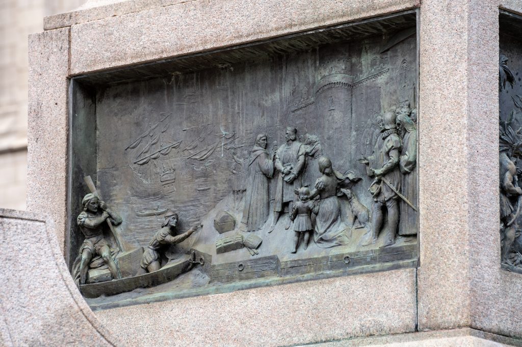 Bronze relief sculpture showing two men moored in a dinghy outside a castle-like structure, with a crowd waiting on the shore. Tall-masted ships are in the harbor behind.