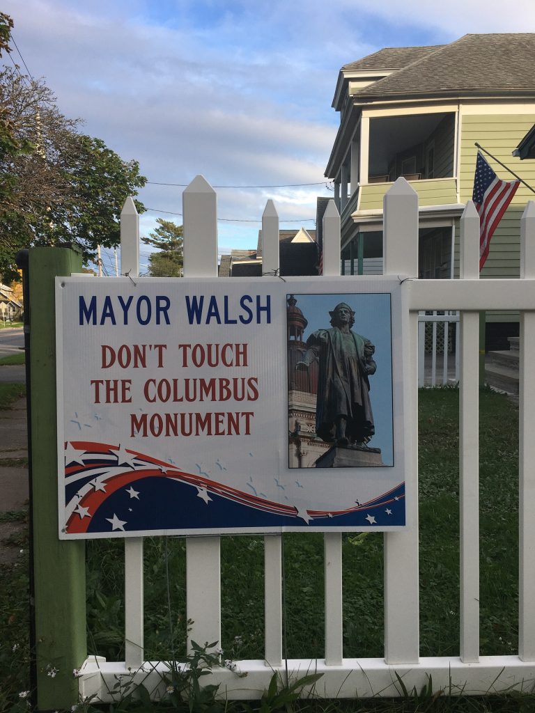 Photograph of a sign on a white picket fence reading "MAYOR WALSH / DON'T TOUCH / THE COLUMBUS / MONUMENT"
