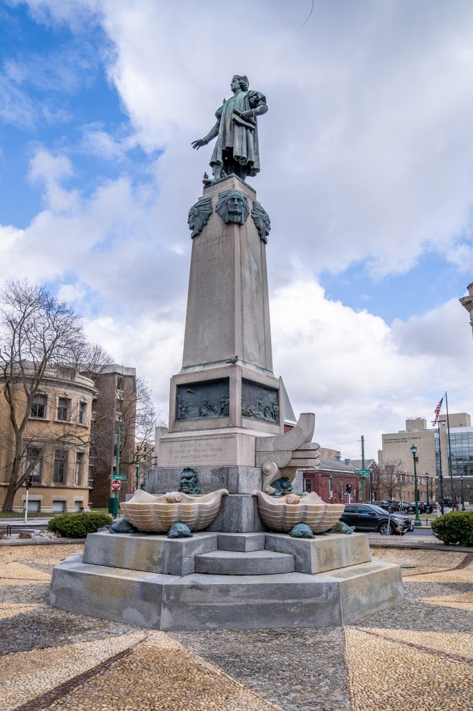 Public sculpture in a park with a bronze statue of Christopher Columbus atop a tall stone pedestal decorated with additional bronze sculptural elements.