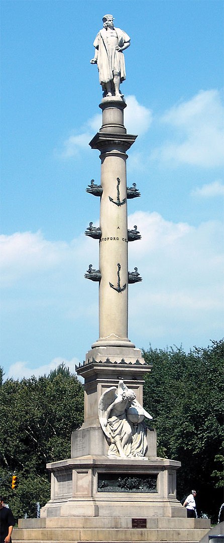 Public monument with a marble statue of Christopher Columbus atop a tale stone pole with smaller sculptures of ships projecting from the sides.
