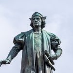 The bronze statue of Christopher Columbus that is at the top of the monument seen in fig. 3. The figure wears a Renaissance-era cap and cloak and carries a rolled chart in his left hand. His right hand is outstretched and his right foot steps forward off the pedestal.