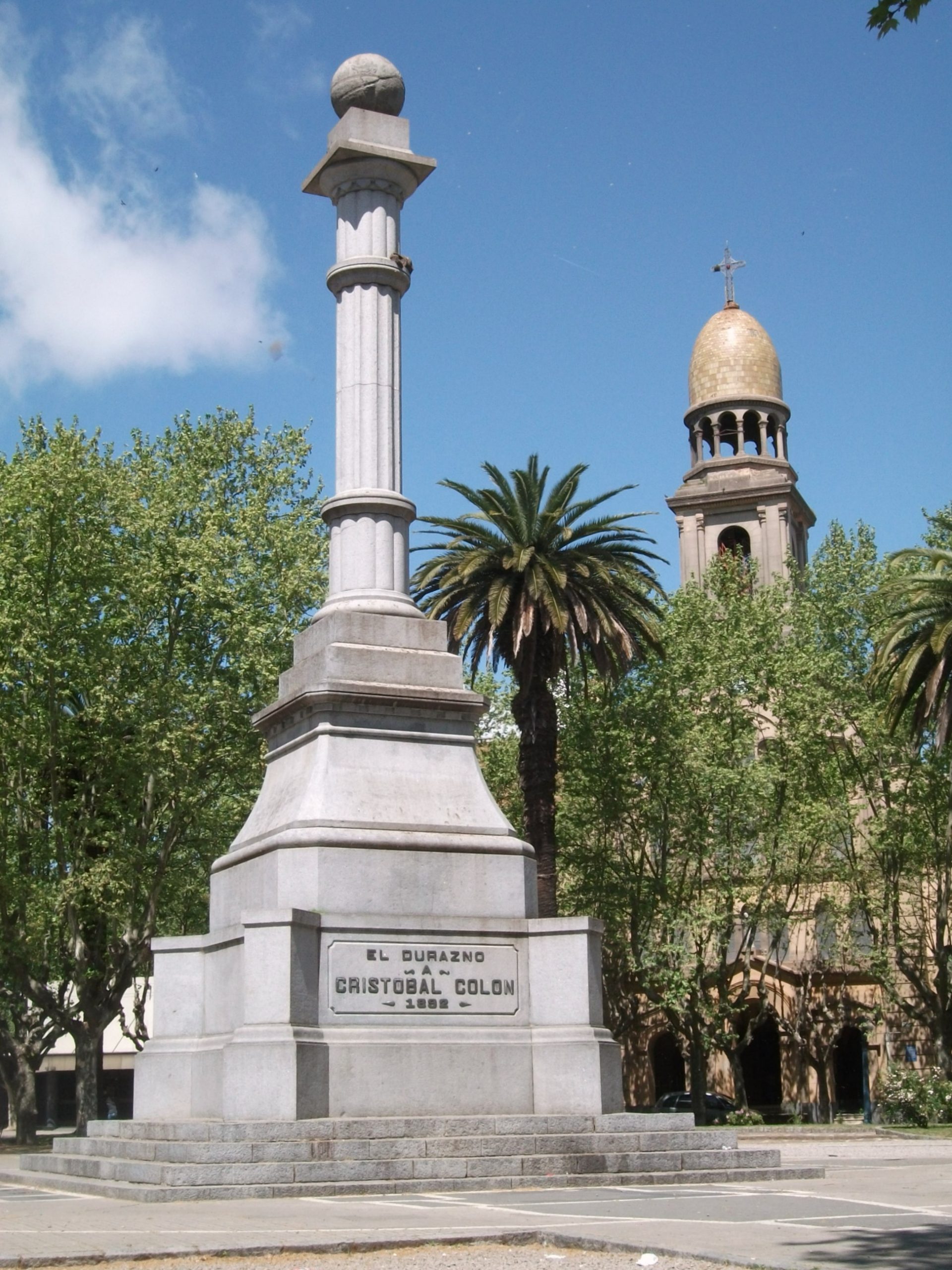 Public monument in a park with palm trees, consisting of a stone column with a globe at its top. On the base an inscription reads "EL DURAZO / A / CRISTOBAL COLON"
