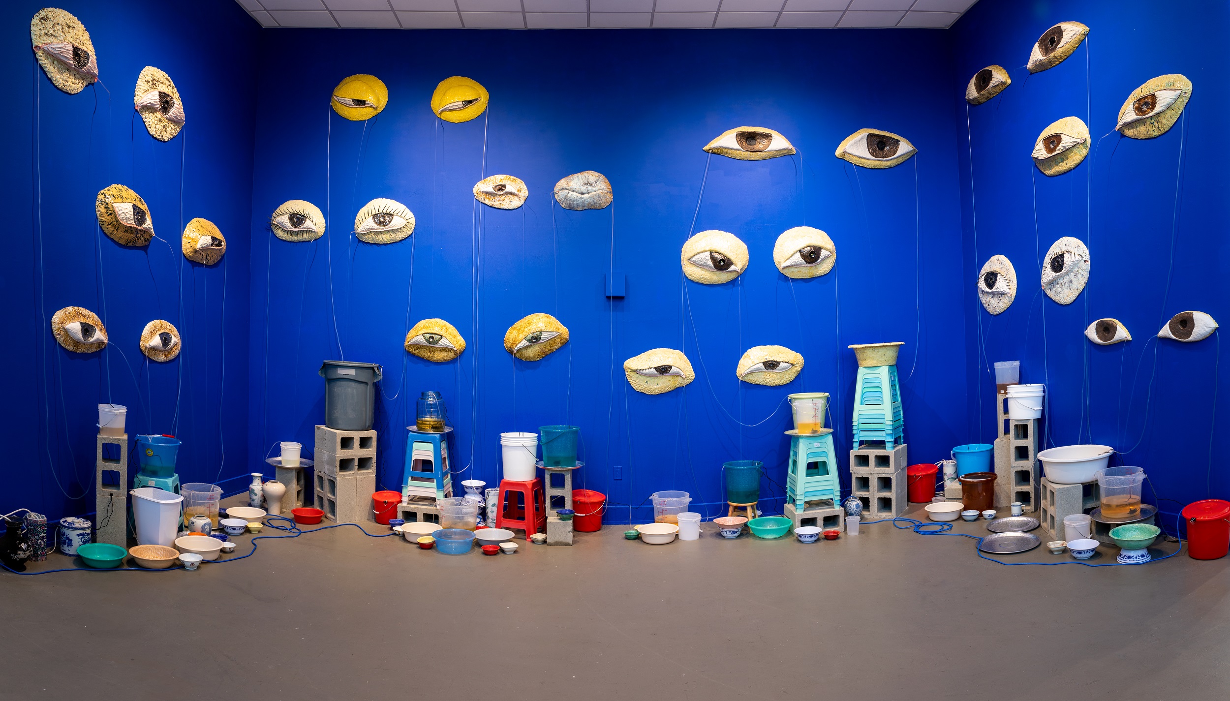 Interior view of a museum gallery with sculptural human eyes hanging on three bright blue walls. On the floor are cinder blocks and various pails, buckets, and dishes.
