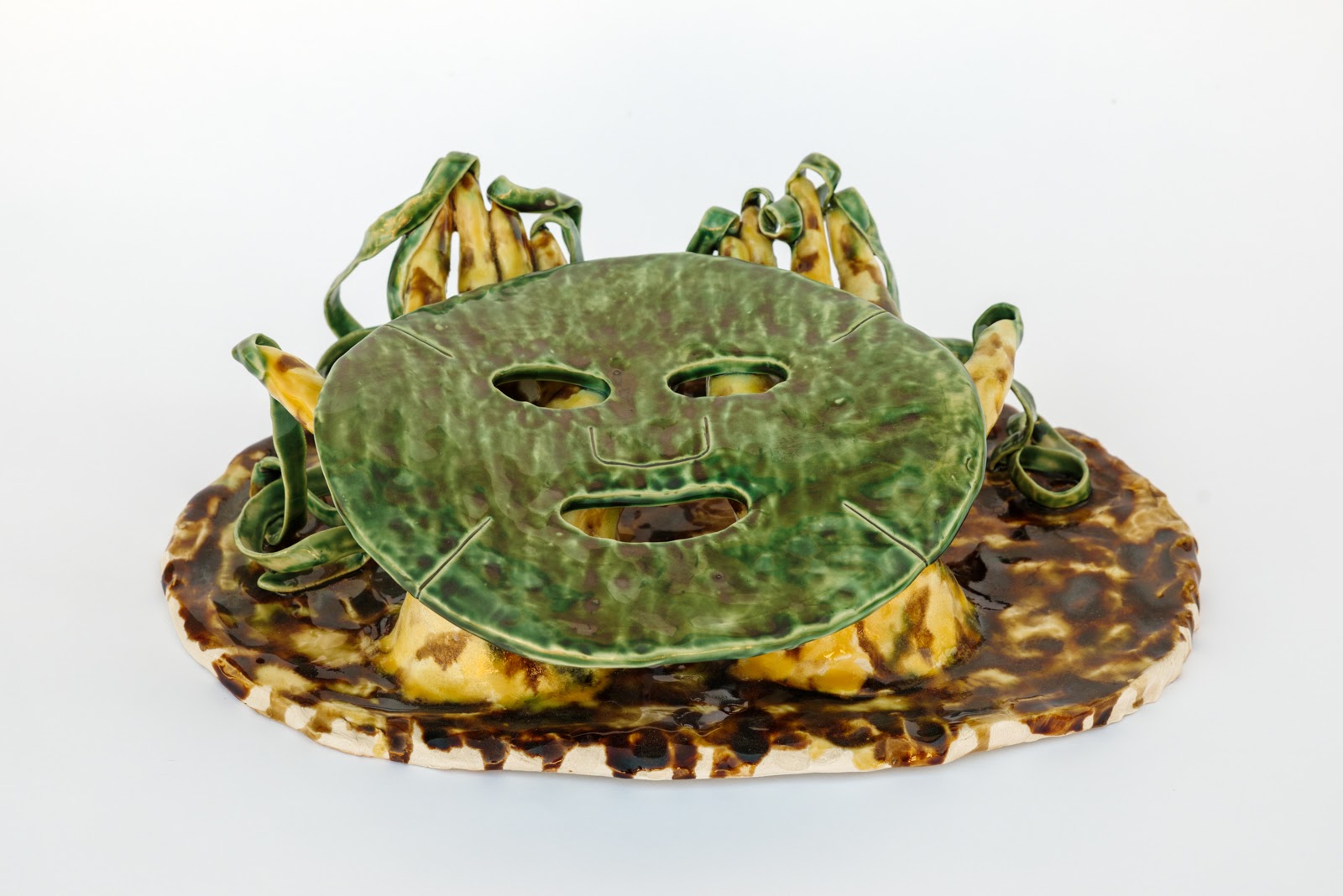 A flat green ceramic face-like shape resting atop another flat ceramic form with brown and yellow glaze.