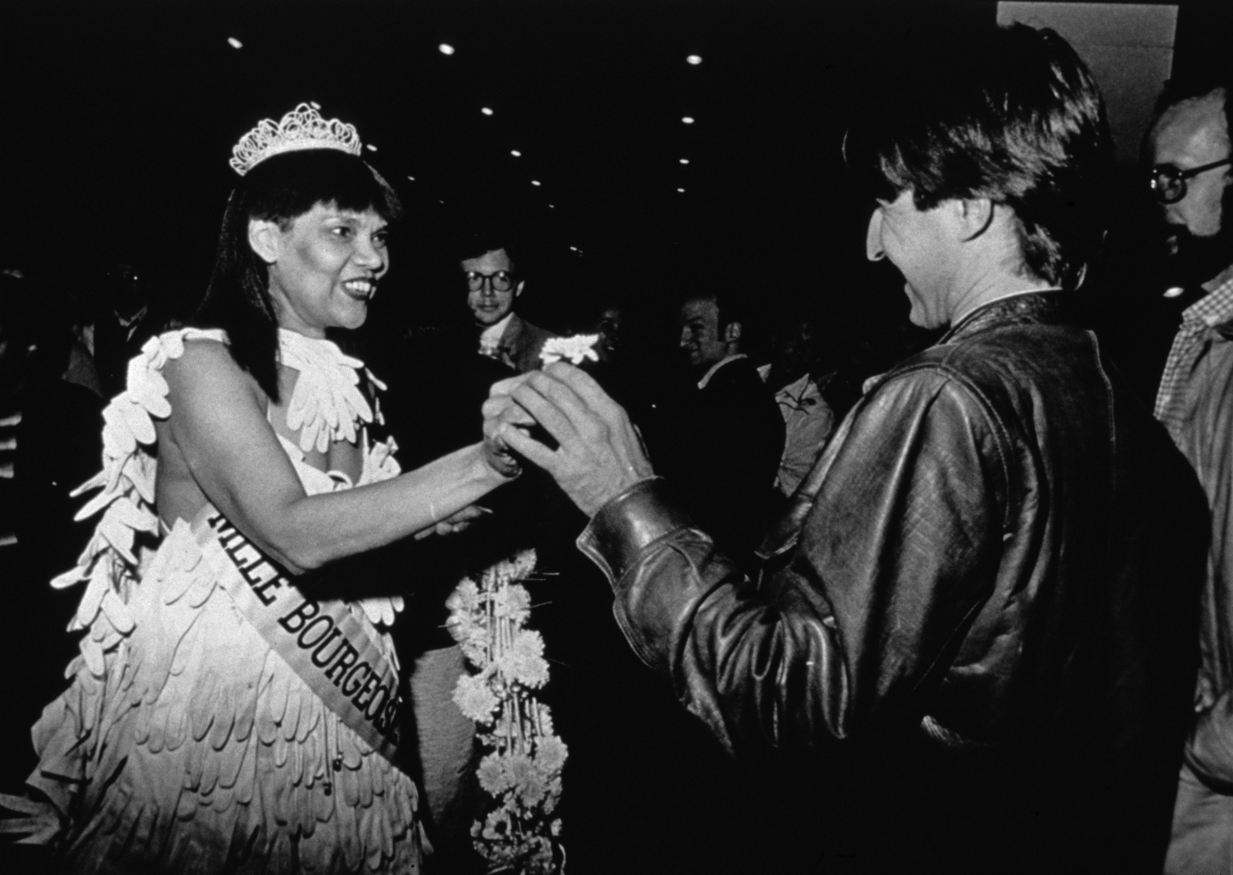 Black-and-white photograph of a Black woman in a tiara, with a pageant sash reading "MLLE BOURGEOSE," smiling and handing a flower to a white man in a leather jacket.