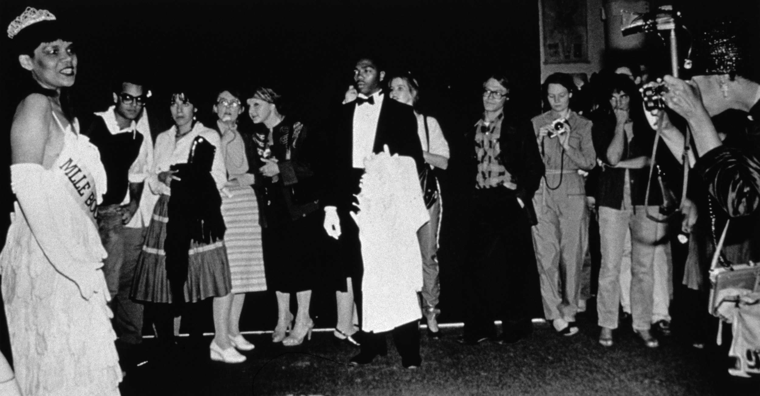 Black and white photograph of a crowd gathered to watch a Black woman in a tiara and a pageant sash. To the far right is a photographer.