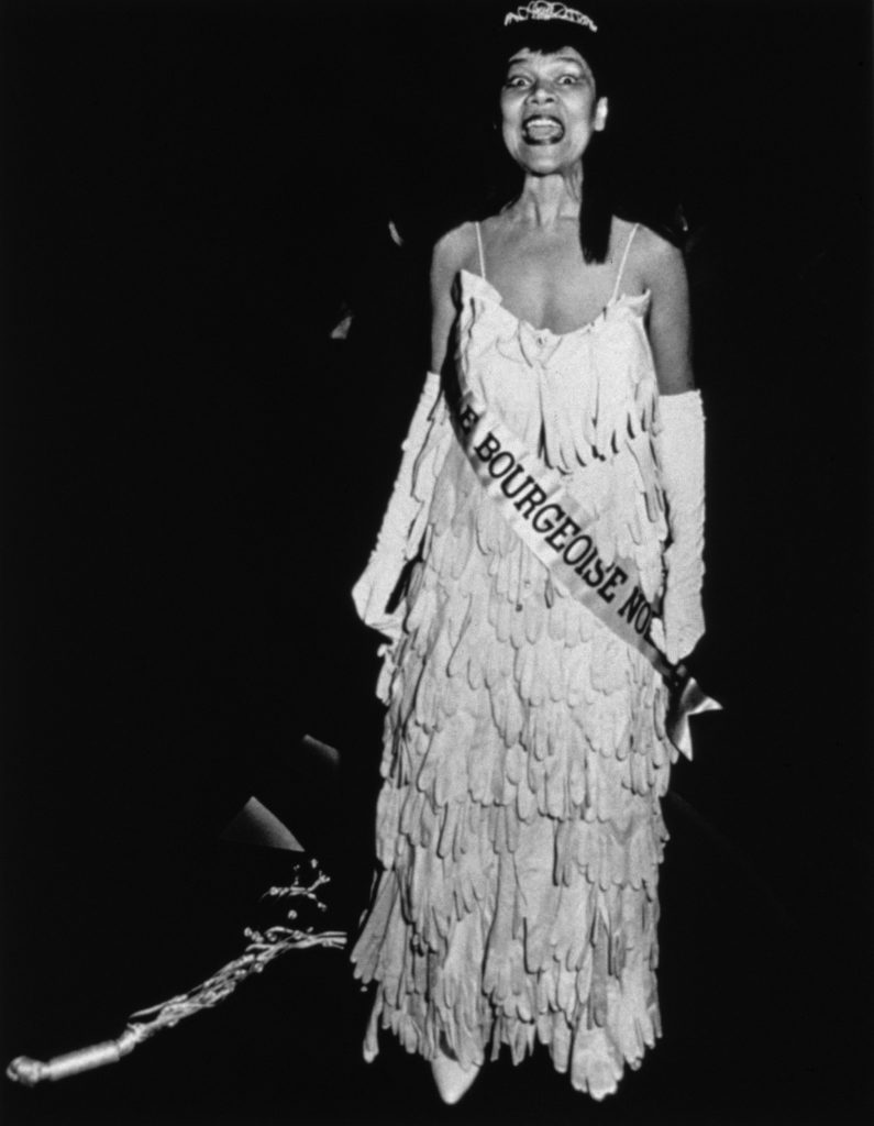 Black-and-white photograph of a Black woman in a tiara, a pageant sash reading "MLLE BOURGEOISE NOIR," and a gown made of white gloves, her eyes and mouth white open as she addresses an unseen audience