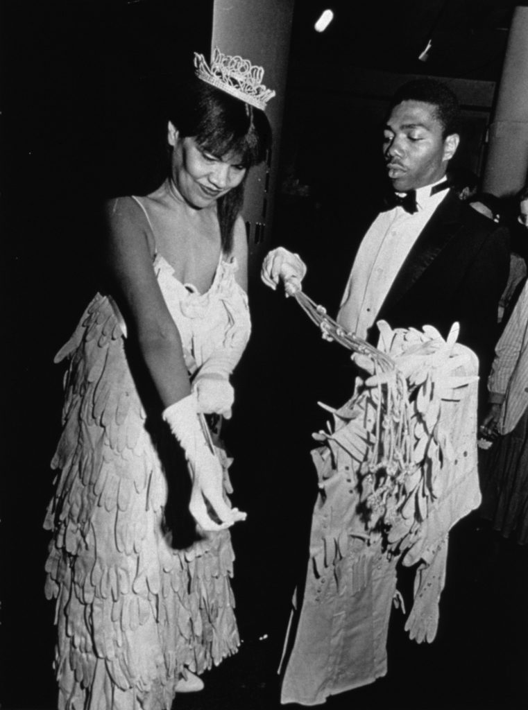 Black-and-white photograph of a Black woman in a tiara and a gown made of gloves removing one glove from her right art; a Black man in a tuxedo hands her a cat-o-nine-tails style whip.