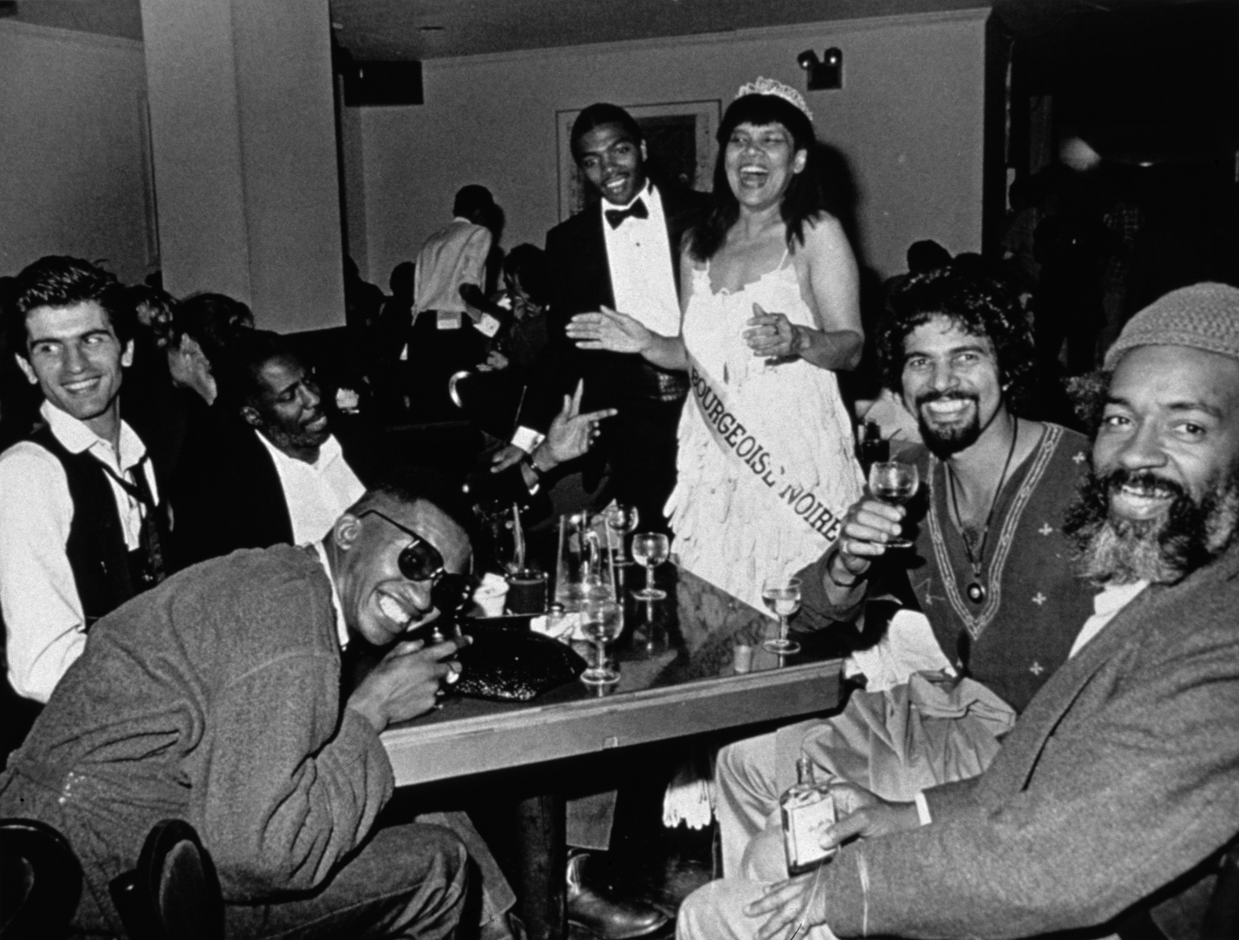 Black-and-white photo of a crowd of people celebrating at a table with drinks in front of them. In the center is a standing Black woman wearing a white dress made of gloves, a tiara, and a pageant sash reading "MLLE BOURGEOISE NOIR."