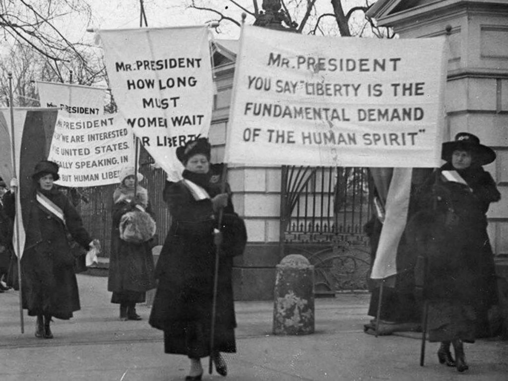 Black-and-white historic photo of women in dark dresses marching in front of a gate, holding signs that read, in part, "MR. PRESIDENT HOW LONG MUST WOMEN WAIT FOR LIBERTY?"